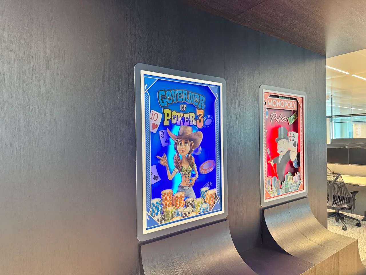 Twee verlichte posters op een donkere muur in een kantoor; één voor Governor of Poker 3 met een cartoonvrouw met cowboyhoed en pokerchips, de andere voor Monopoly Poker met de Monopoly-man die pokerkaarten vasthoudt.