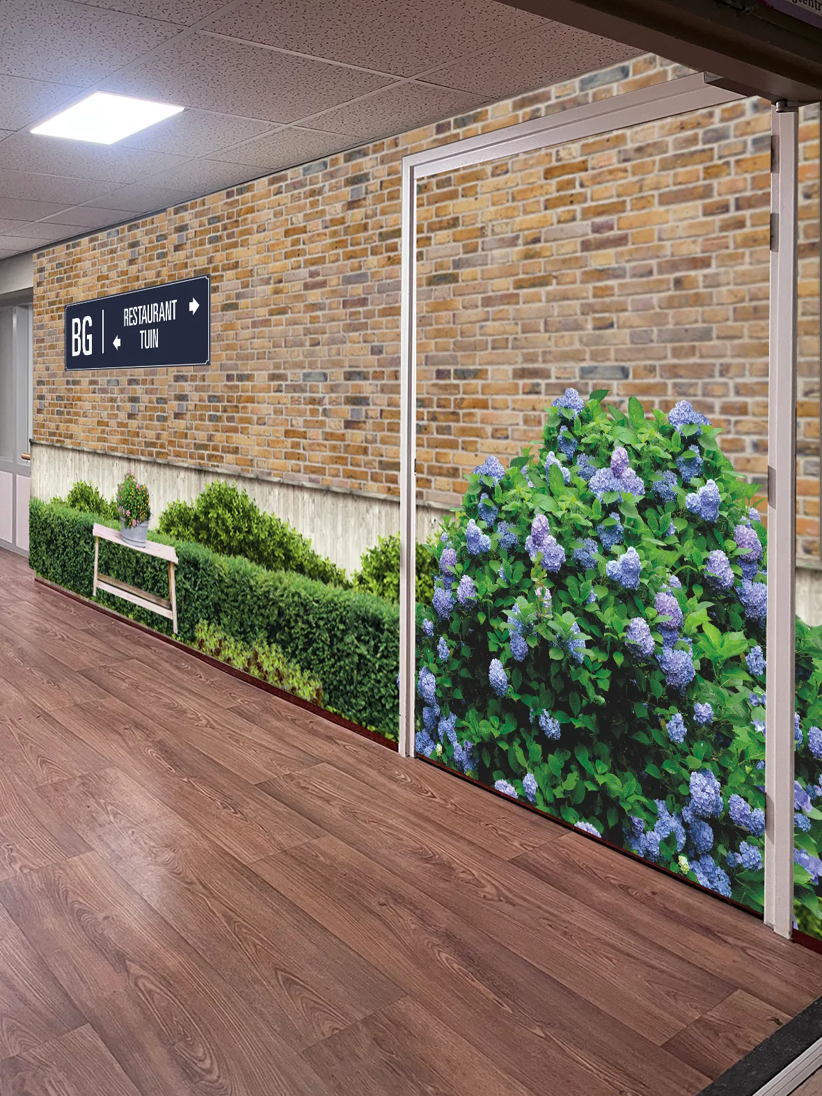 Interieurgang met houten vloer en baksteenbehang met een tuinscène van groene struiken en paarse bloemen, met een bord dat de weg naar het restaurant en de tuin aangeeft.