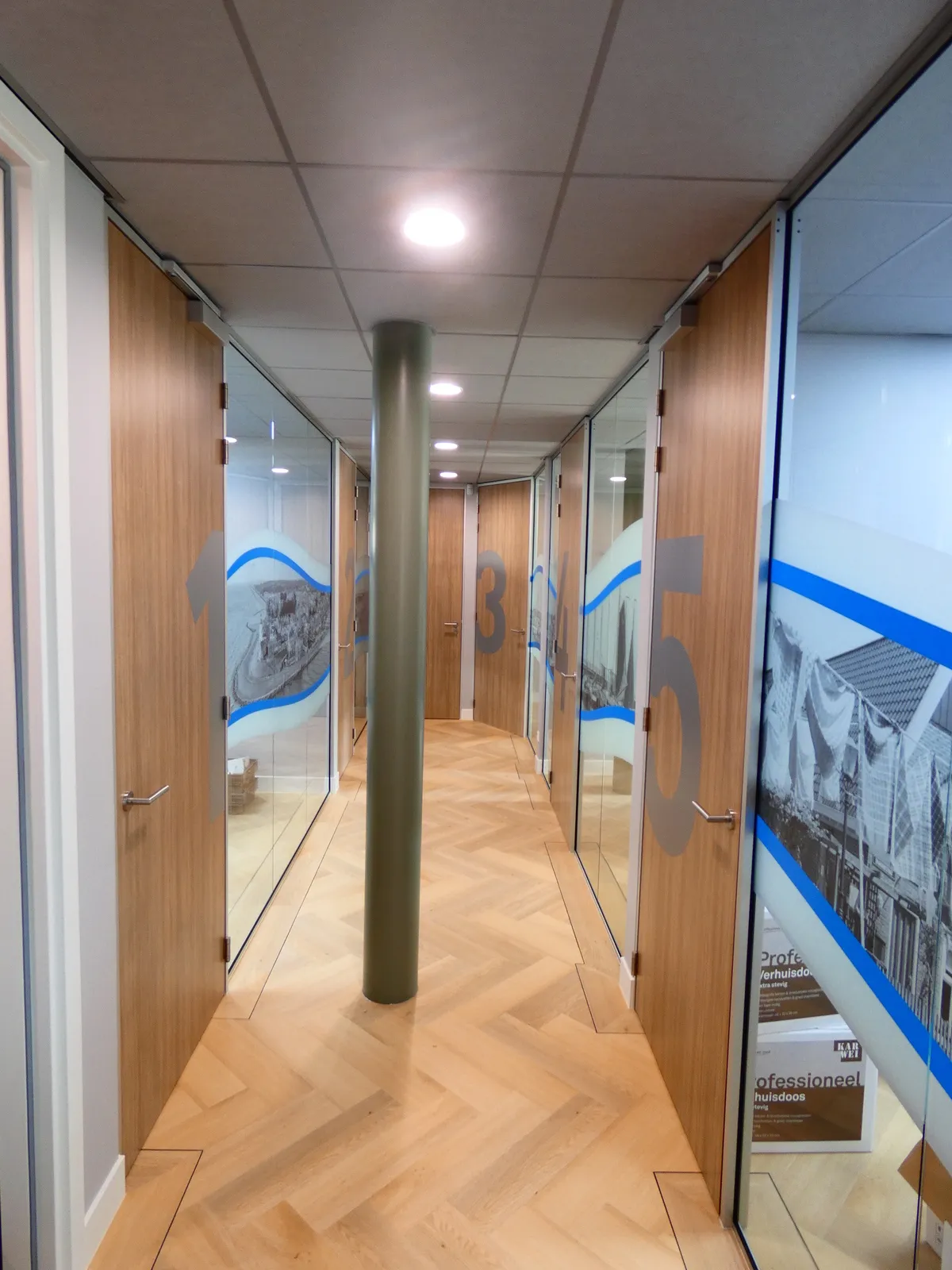 Gang met houten deuren genummerd 1 tot 5 en glazen wanden met blauw golfpatroon en monochrome afbeeldingen.