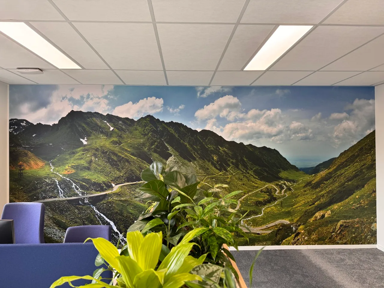 Kantoorinterieur met paarse stoelen, groene planten en een grote muurprint van een kronkelende bergweg onder een deels bewolkte lucht.