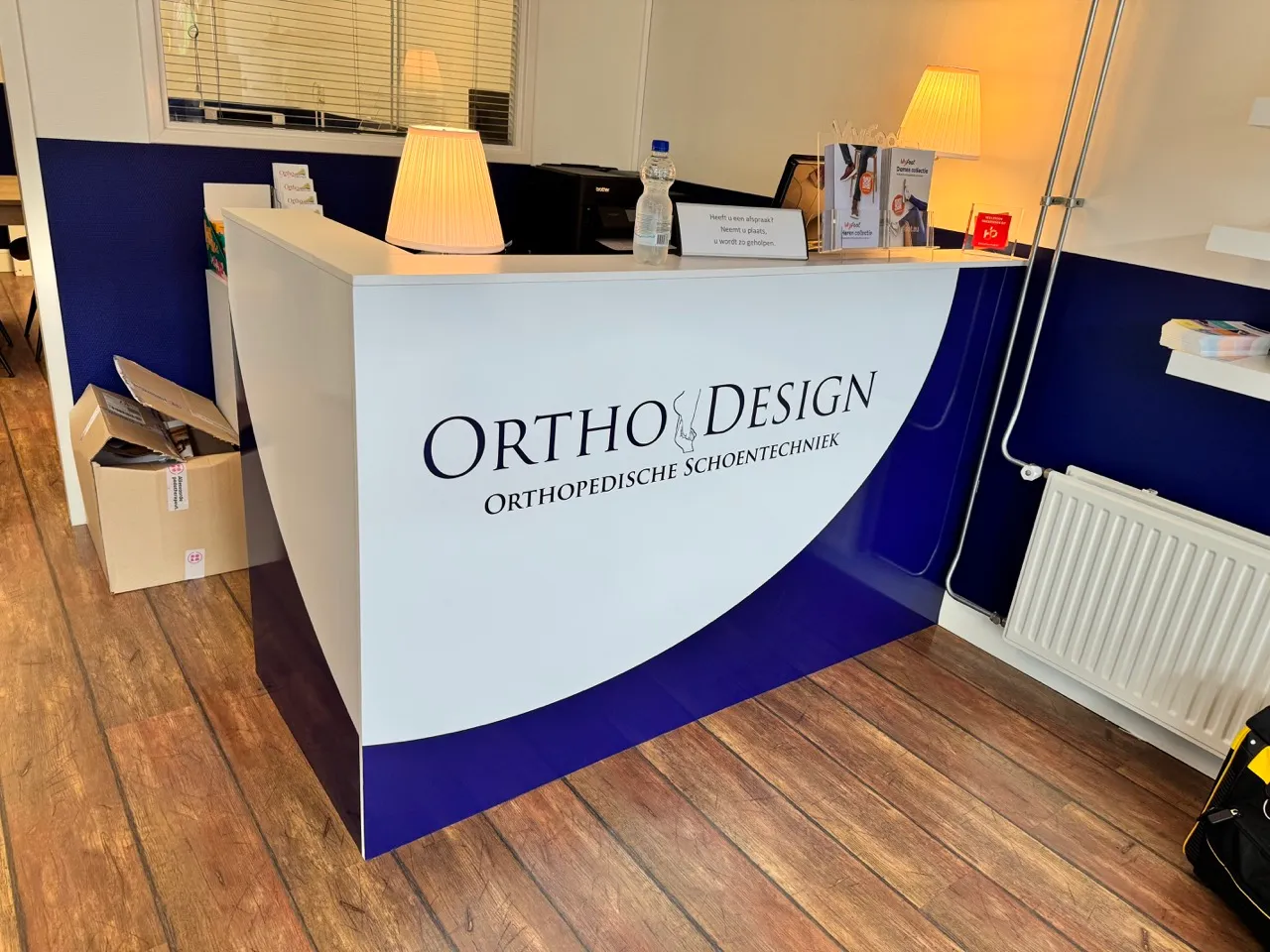 Balie met bord ‘Ortho Design Orthopedische Schoentechniek’ in een kliniek, met twee lampen, dozen en brochures op de balie.