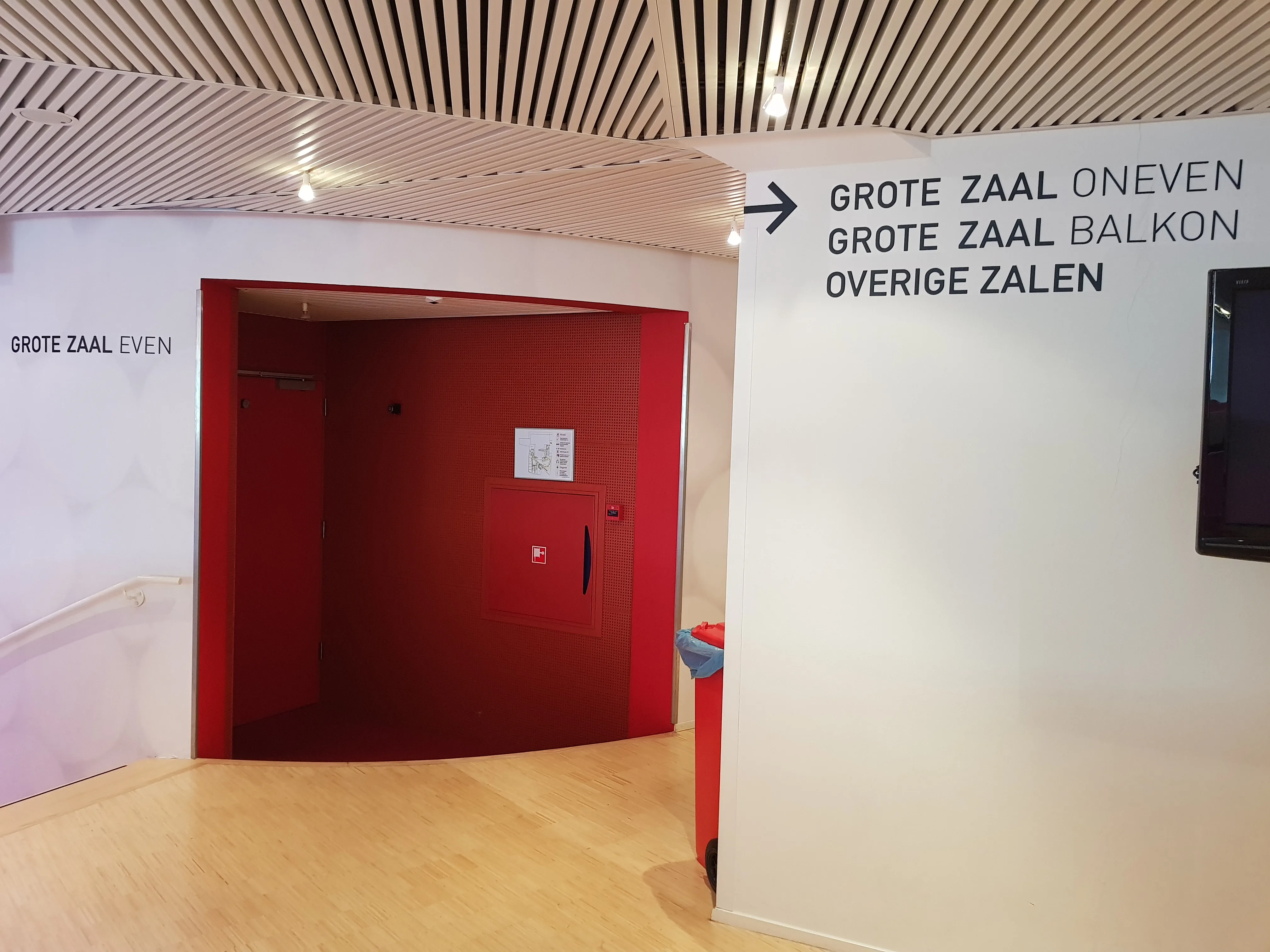 Interieurgang met witte muren en houten vloer, een rode deuropening en richtingborden in het Nederlands naar verschillende zalen, waaronder Grote Zaal en Overige Zalen.