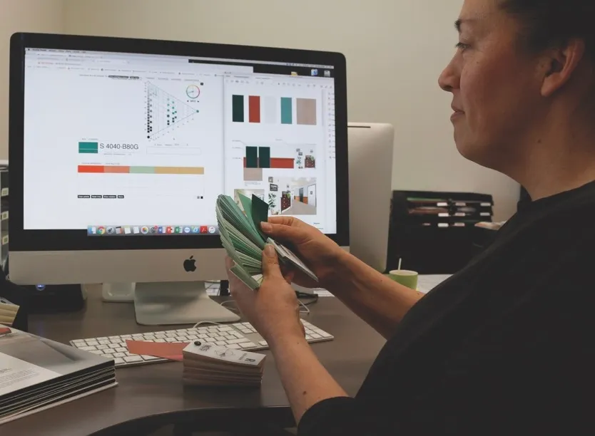 Persoon houdt een waaier met groene kleurstalen voor een computerscherm waarop een kleurenpalet en designsoftware te zien zijn.