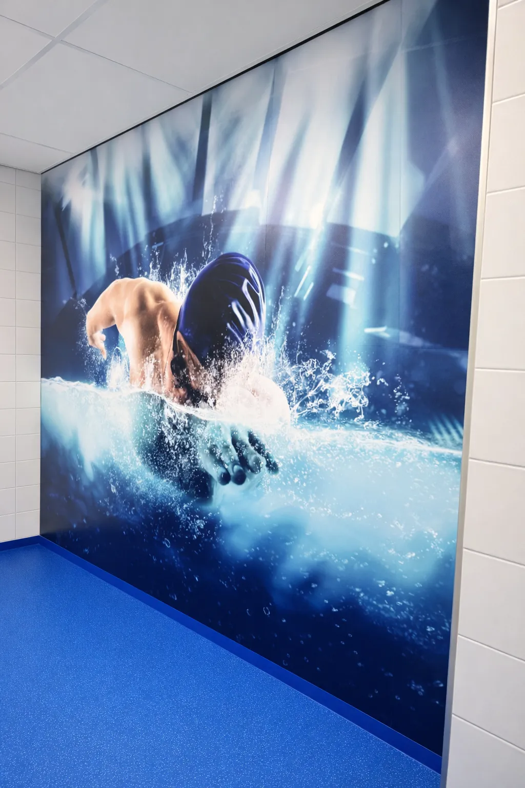 Muurschildering van een zwemmer met badmuts die spetters in het water maakt, geplaatst op een blauwe vloer met witte betegelde wanden eromheen.