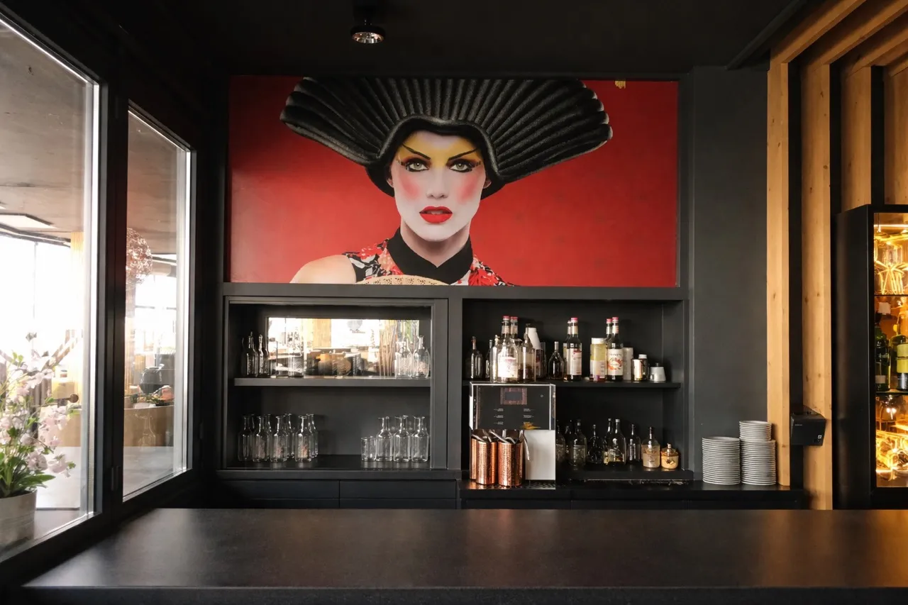 Modern barinterieur met een groot portret van een vrouw met dramatische make-up en een zwarte waaiervormige hoed boven glazen planken met flessen en glazen.