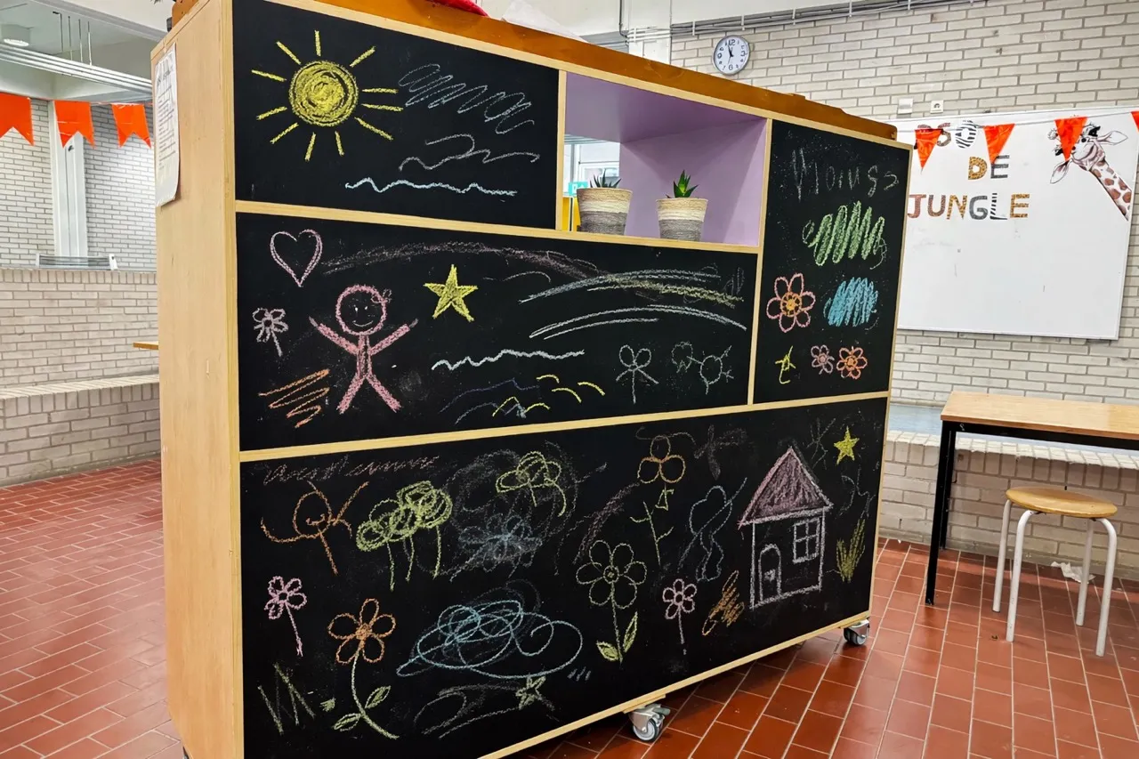 Houten verrijdbaar bord met meerdere krijtborden met kleurrijke krijttekeningen van een zon, stickfiguren, harten, sterren, bloemen en een huis in een helder verlicht klaslokaal.