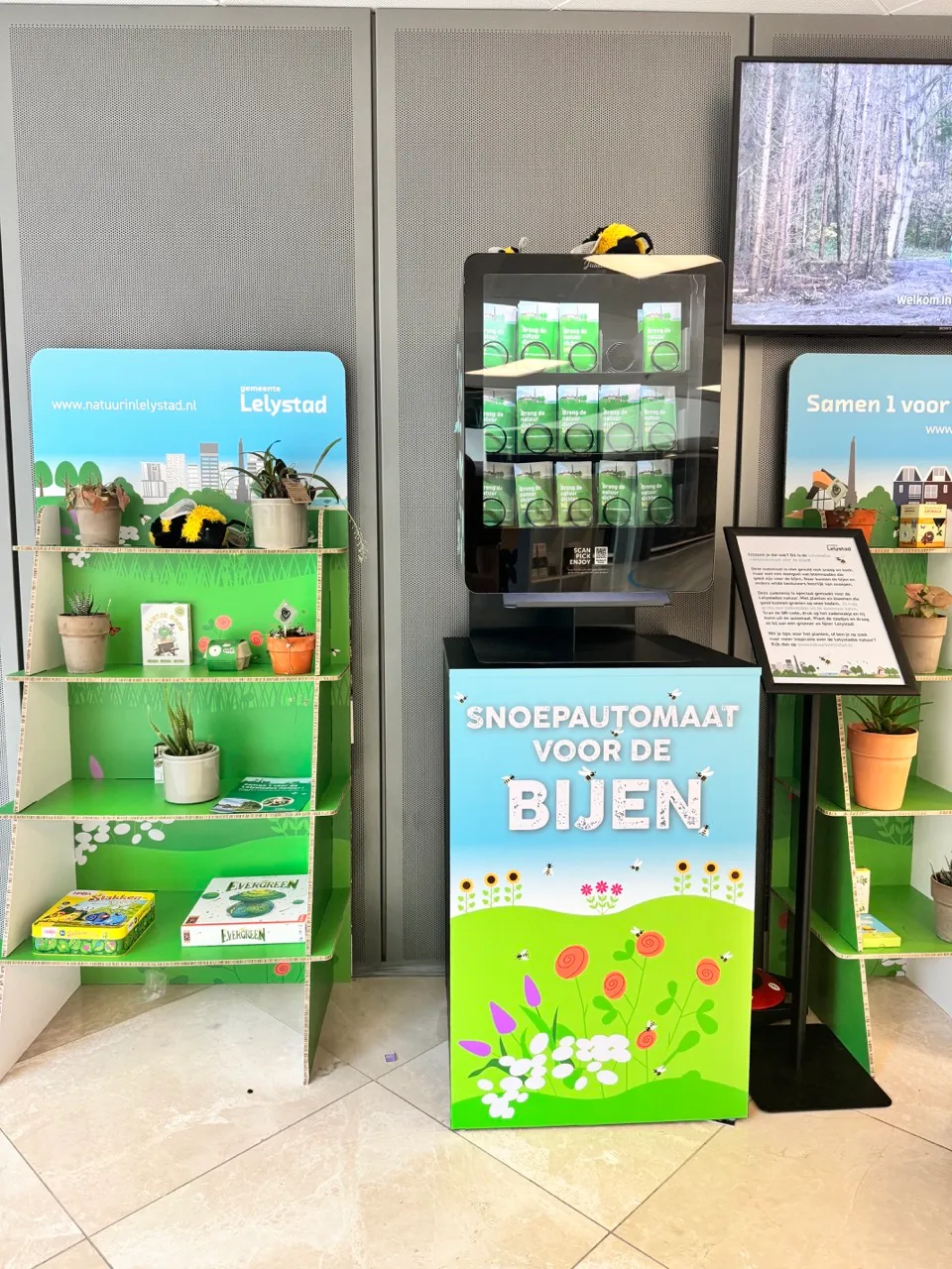 Binnenpresentatie met een ‘candy machine for bees’ automaat gevuld met groene doosjes, omringd door kartonnen schappen met planten in potten en informatiemateriaal.