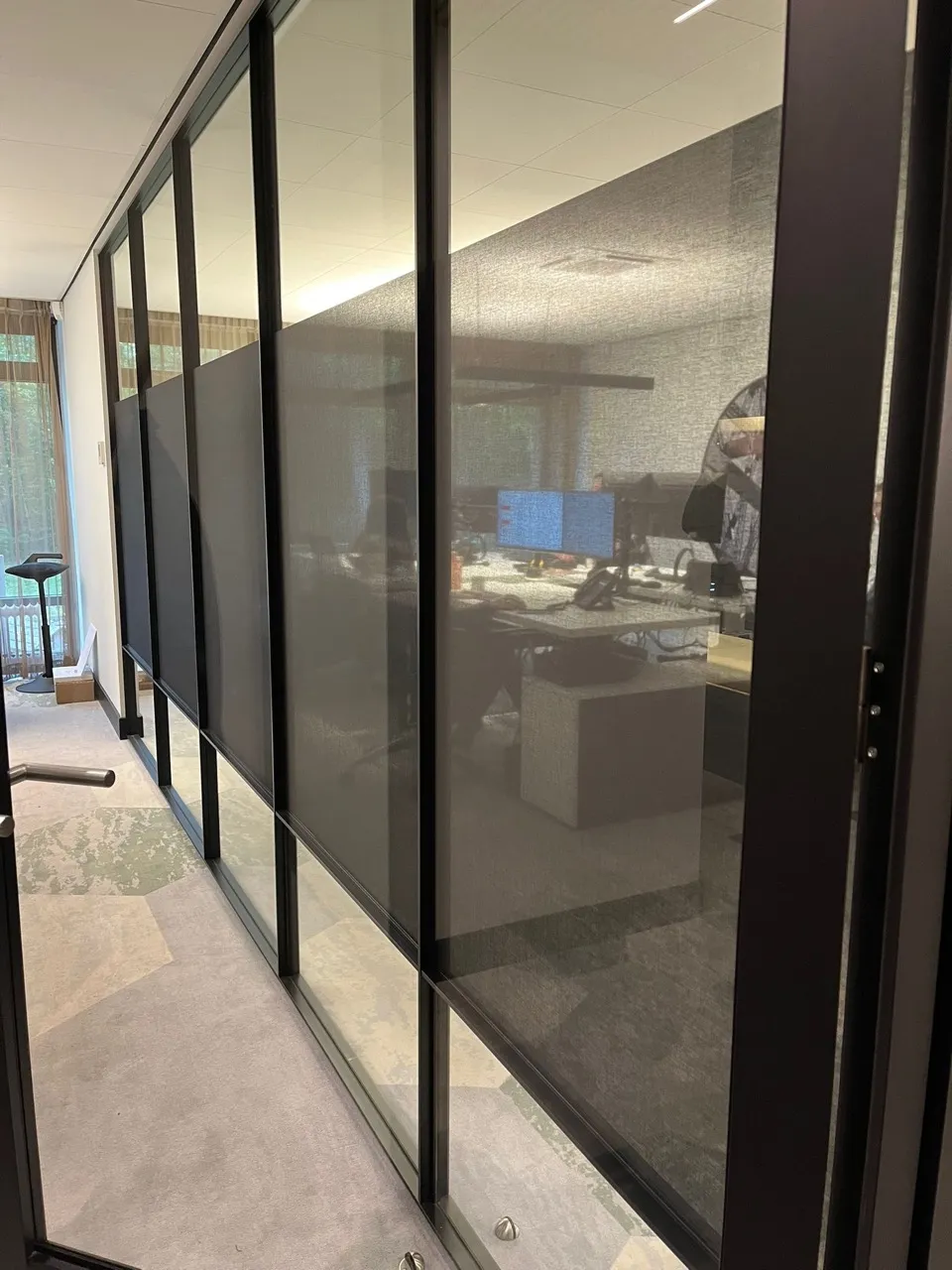 Kantoorgang met glazen wanden gedeeltelijk bedekt met donkere stoffen jaloezieën, met zicht op bureaus en computerschermen binnen.