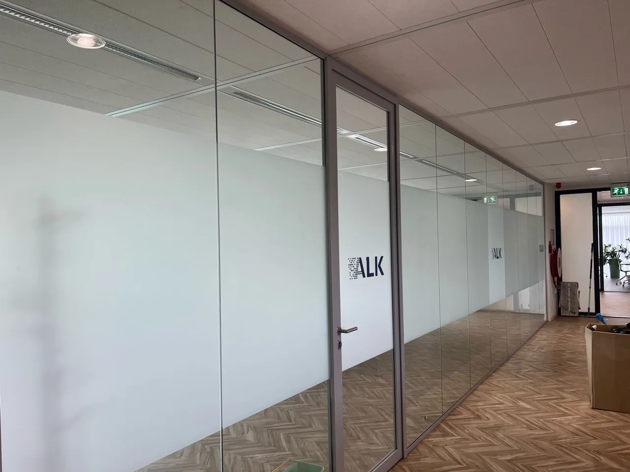 Moderne kantoorgang met glazen wanden en een deur met het ALK-logo, parketvloer en plafondverlichting.