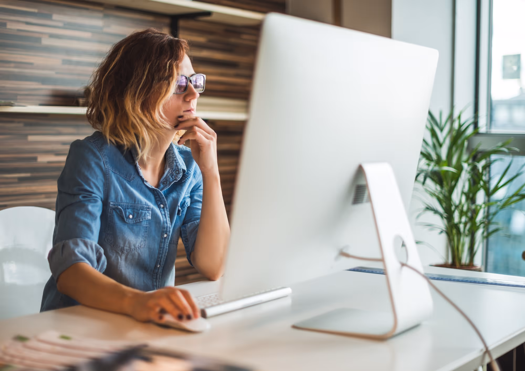 Woman wearing glasses and a denim shirt sitting at a desk, using a computer mouse while looking thoughtfully at a large desktop monitor.