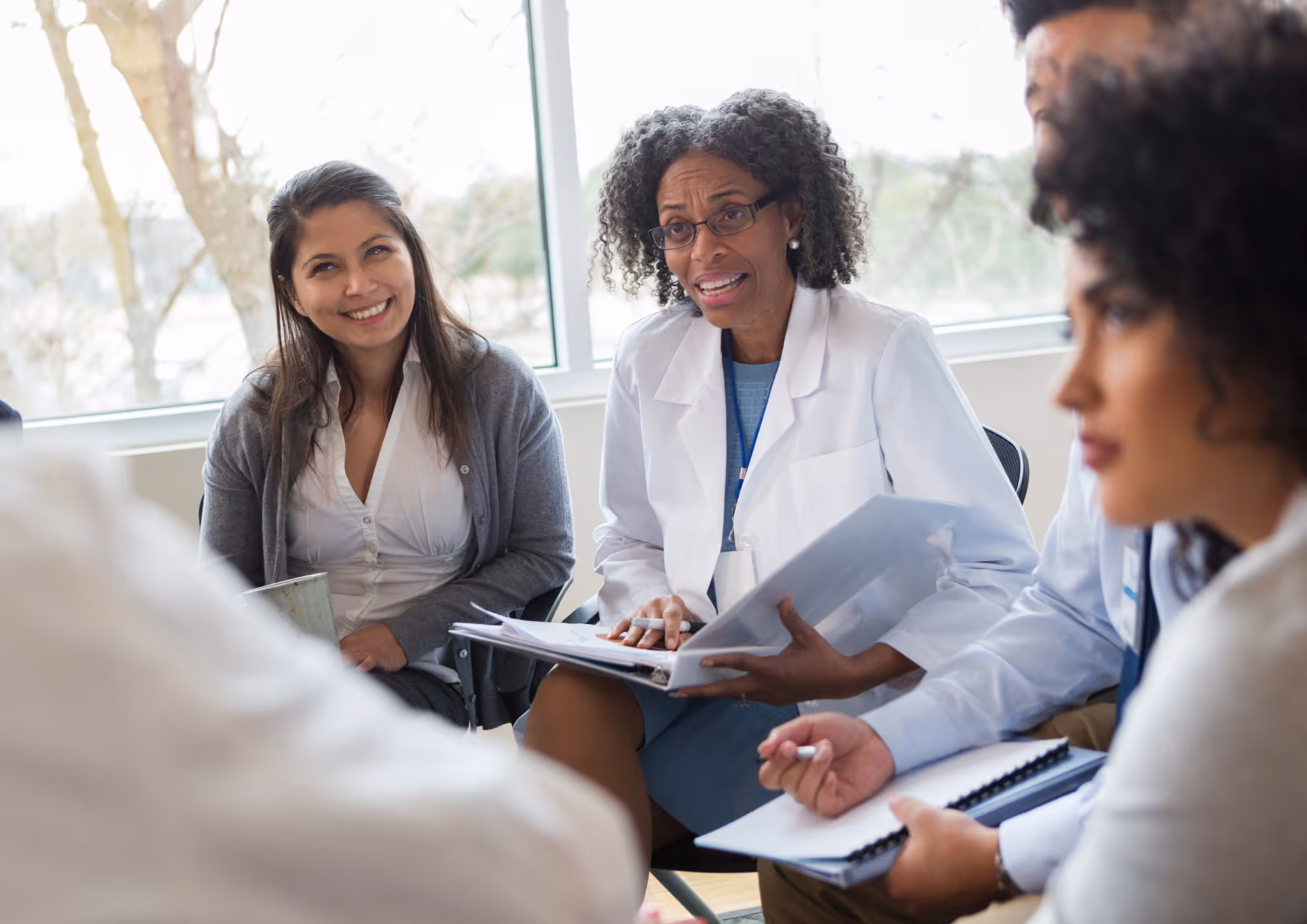 Diverse group of professionals in a meeting, with one woman in a white lab coat speaking and others listening attentively.