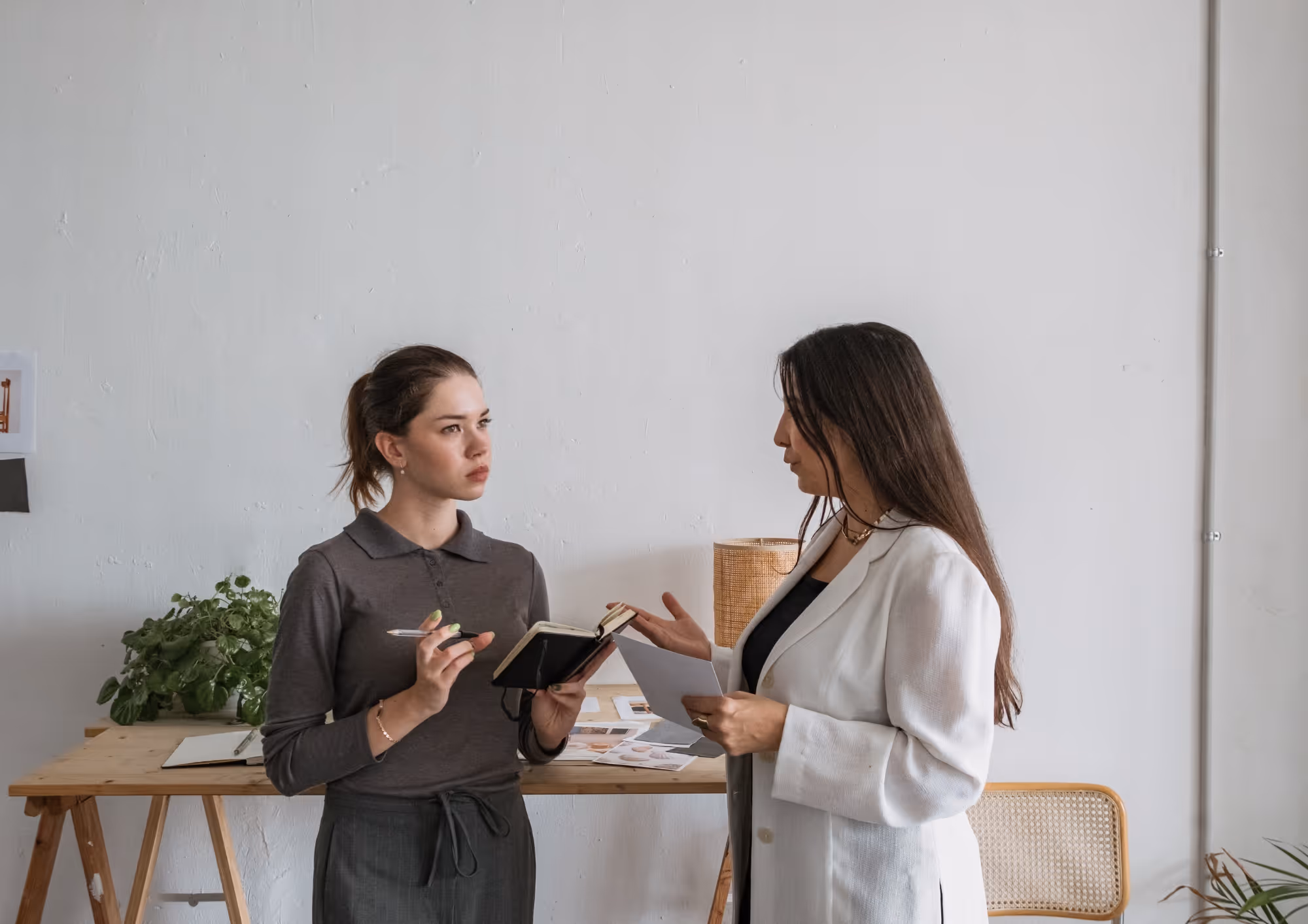 Two women in a minimalist office having a discussion, one holding a pen and notebook, the other holding a document.