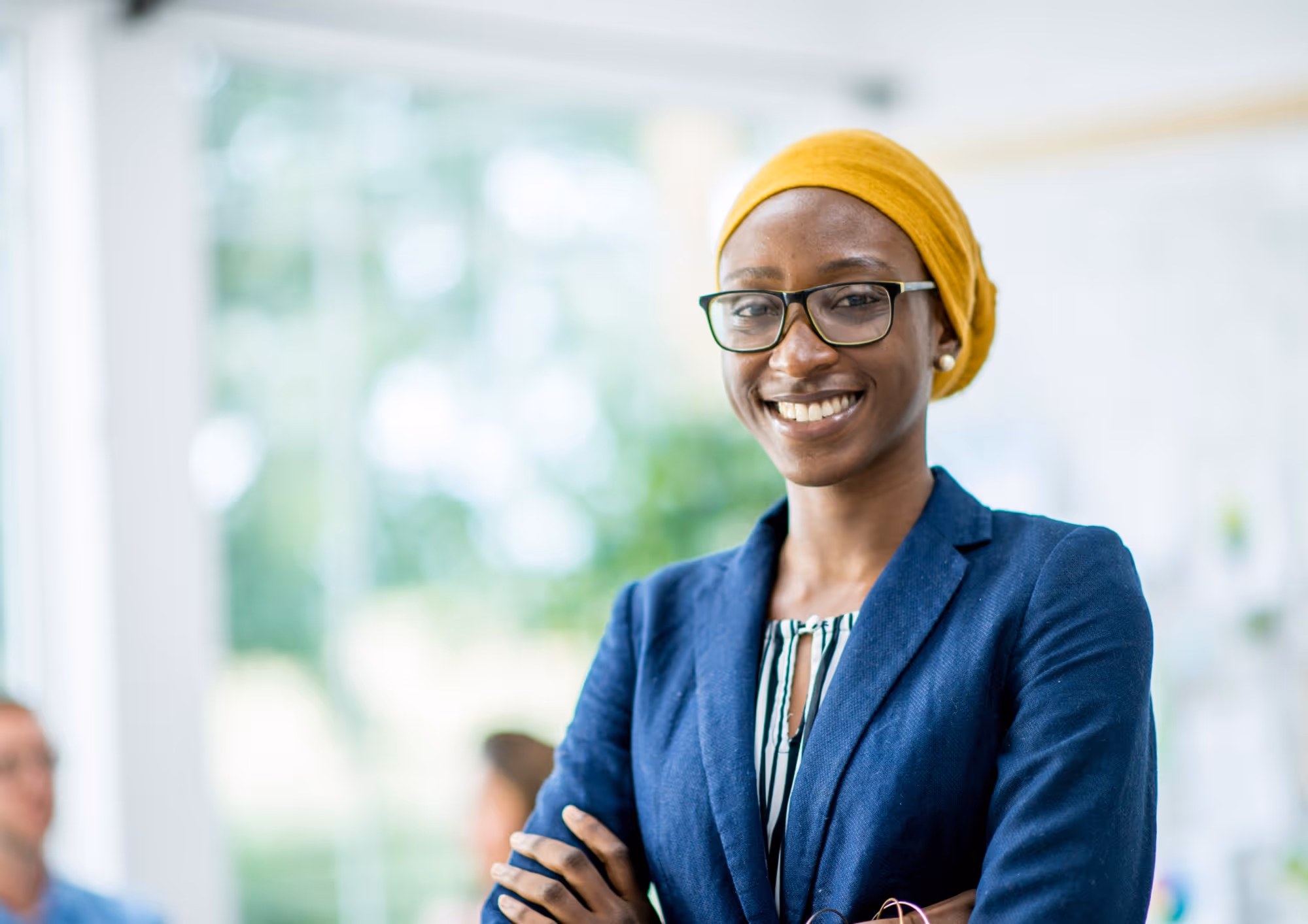 Smiling woman wearing glasses and a yellow headscarf stands with crossed arms in a bright office.