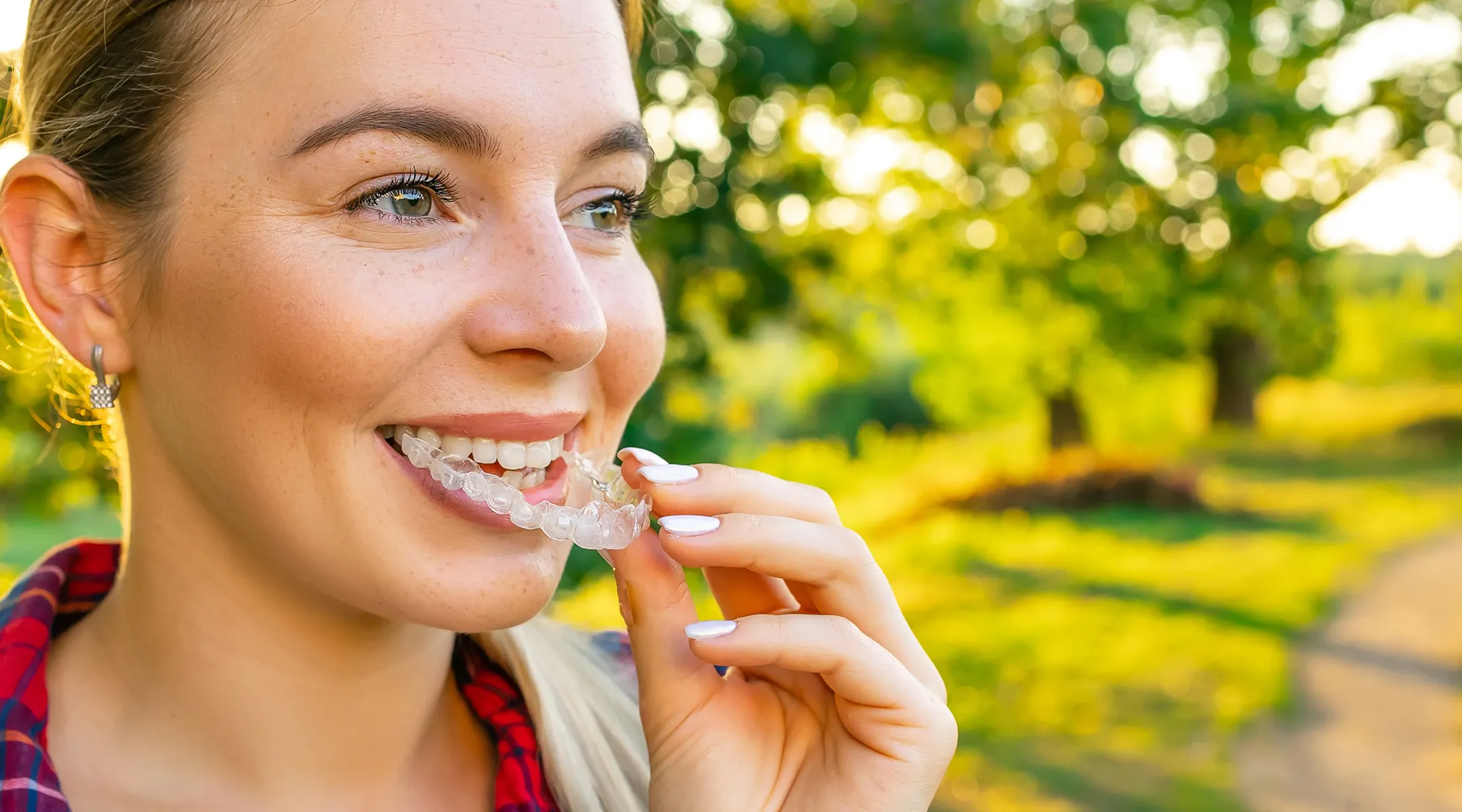 Woman outdoors holding a clear dental aligner near her mouth with a smile.