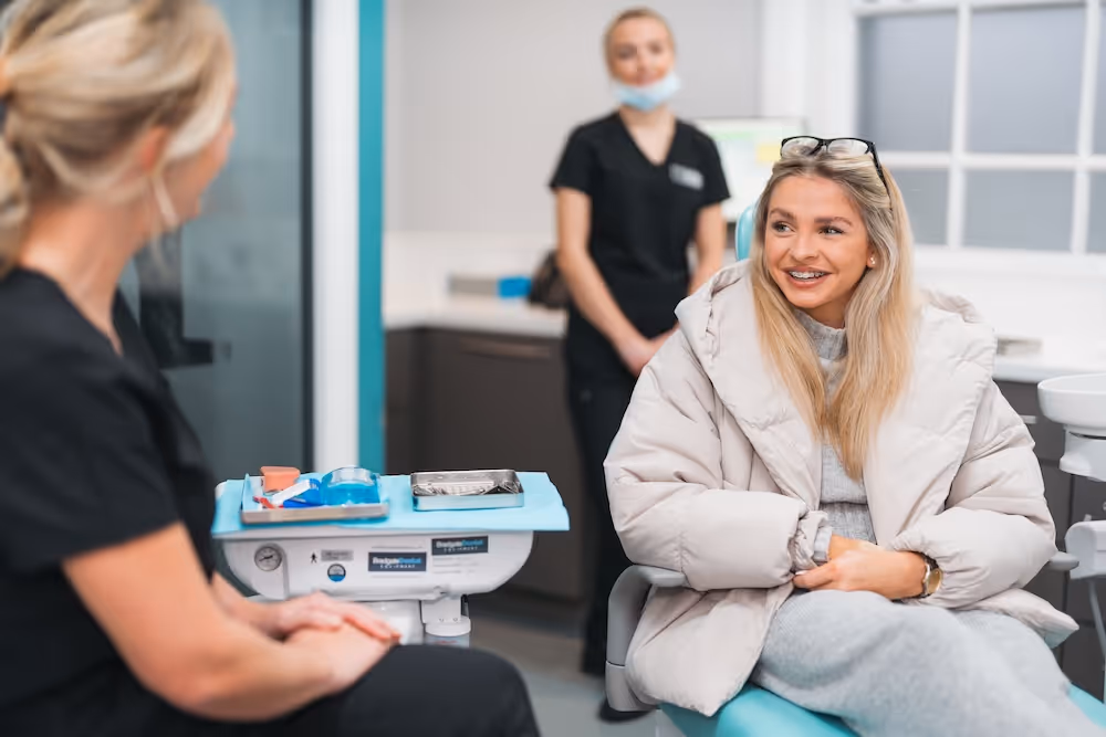 Young woman wearing a beige puffer jacket sitting in a dental chair, smiling and talking to a female dental professional in black scrubs.