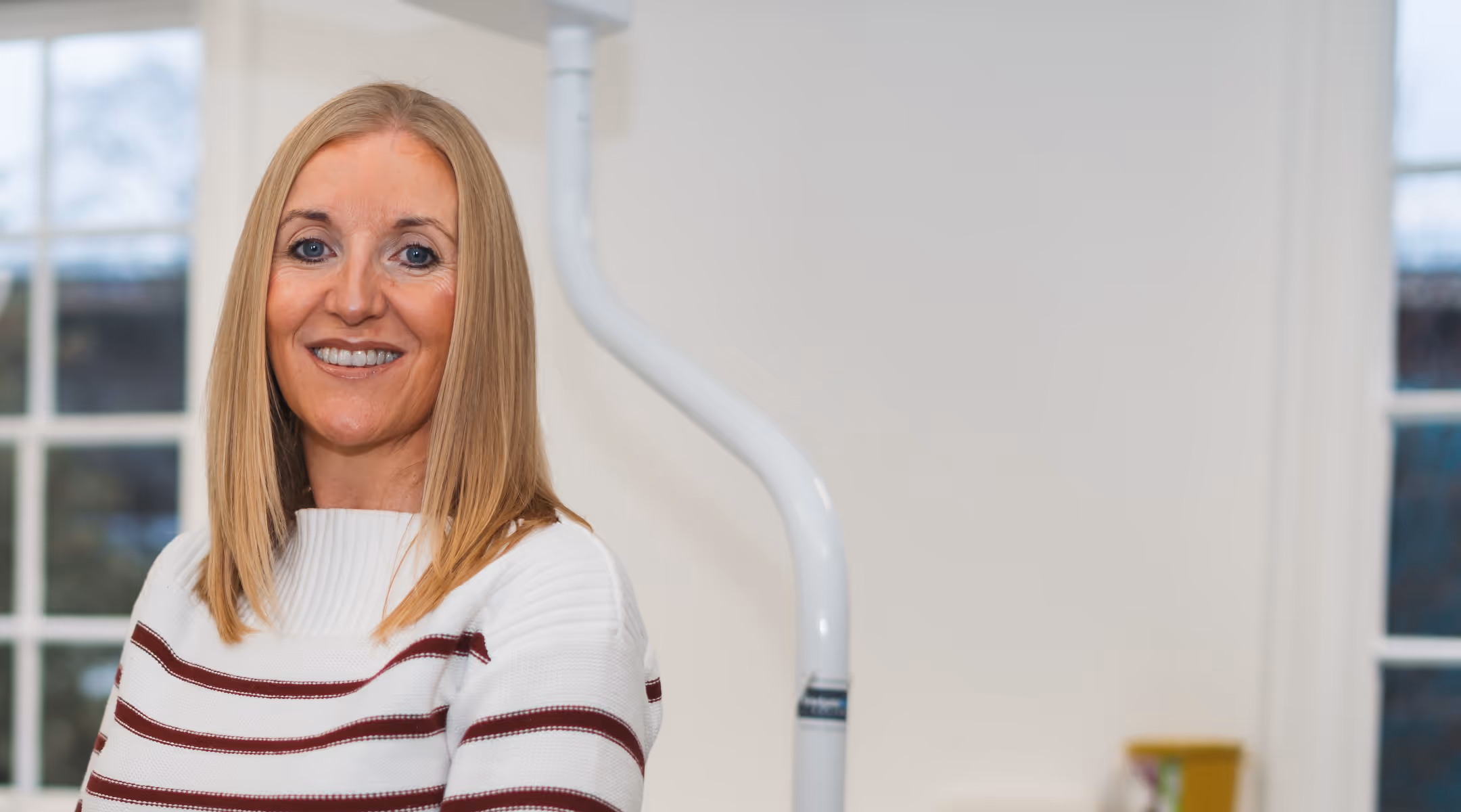 Smiling middle-aged woman with straight blonde hair wearing a white sweater with maroon stripes in a bright room.