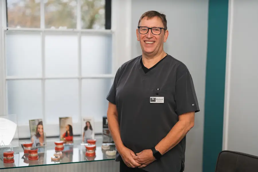 Smiling male dental professional wearing glasses and dark scrubs standing beside a glass table with dental models and brochures.