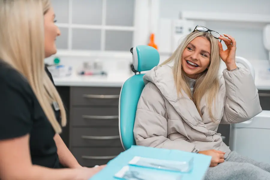 Smiling young woman with braces sitting in a dental chair talking to a female dental professional.