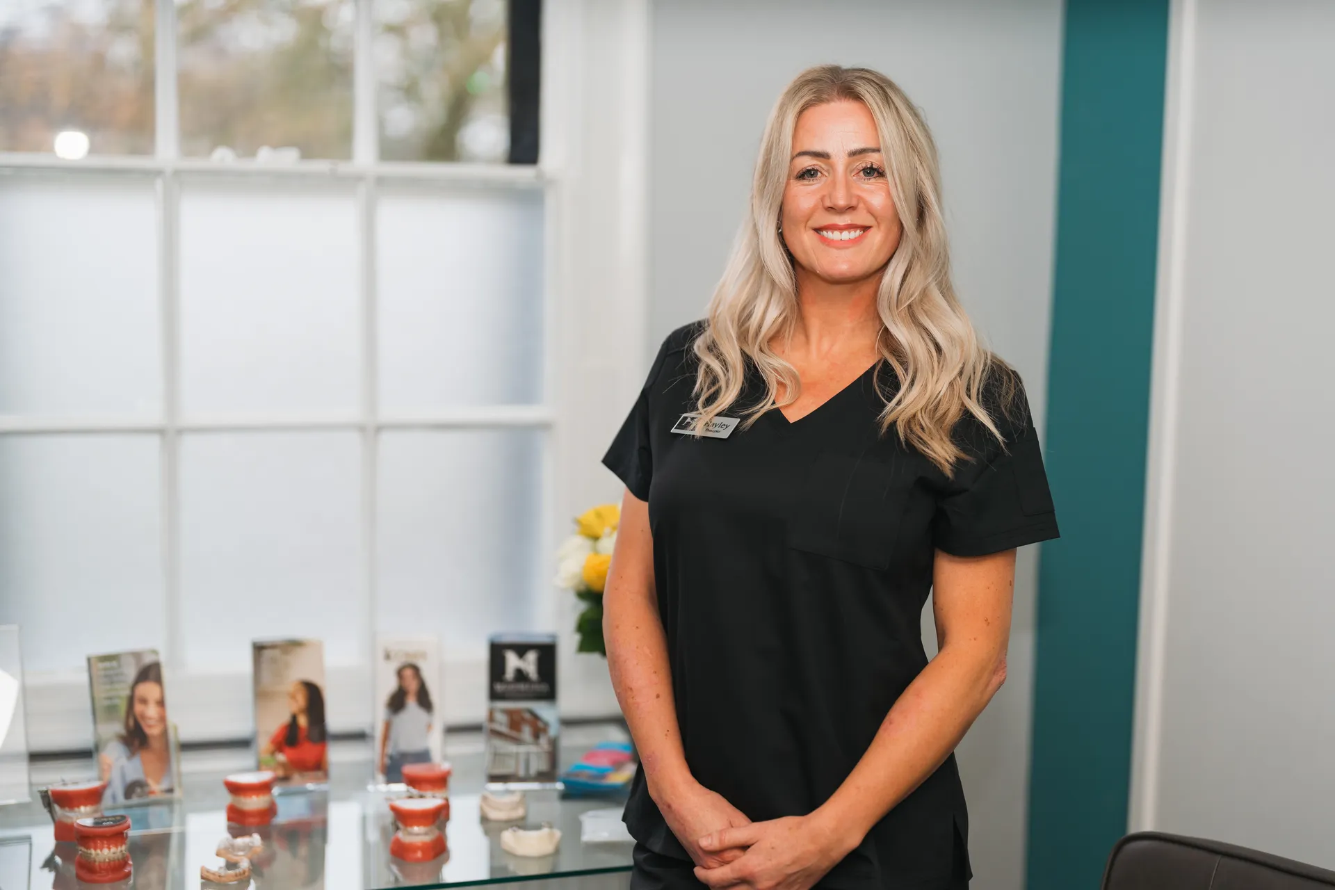 Smiling blonde woman in black scrubs standing in a dental office with dental molds on a glass table.