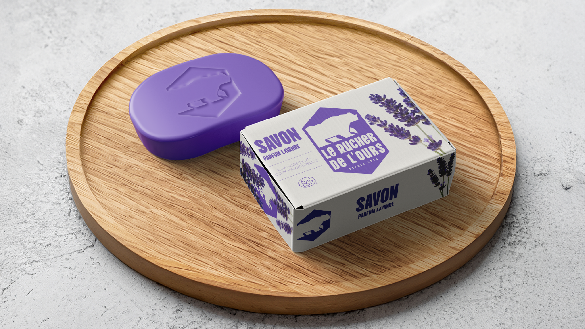 Purple lavender-scented soap bar with a bear logo next to its matching box on a round wooden tray.