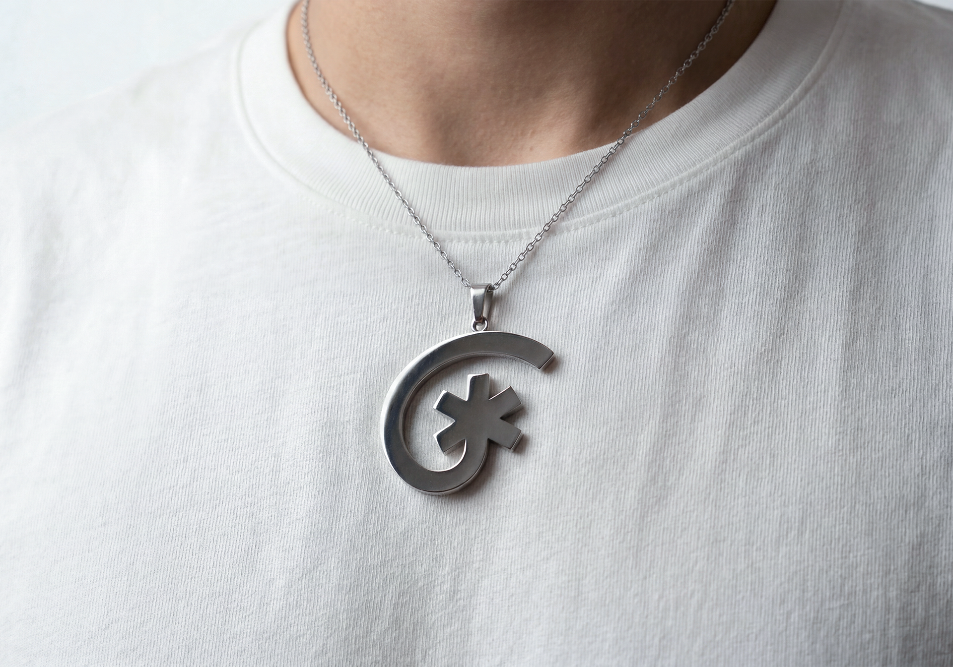 Collier avec un pendentif en métal argenté en forme de demi-cercle avec un astérisque au centre, porté sur un t-shirt blanc.