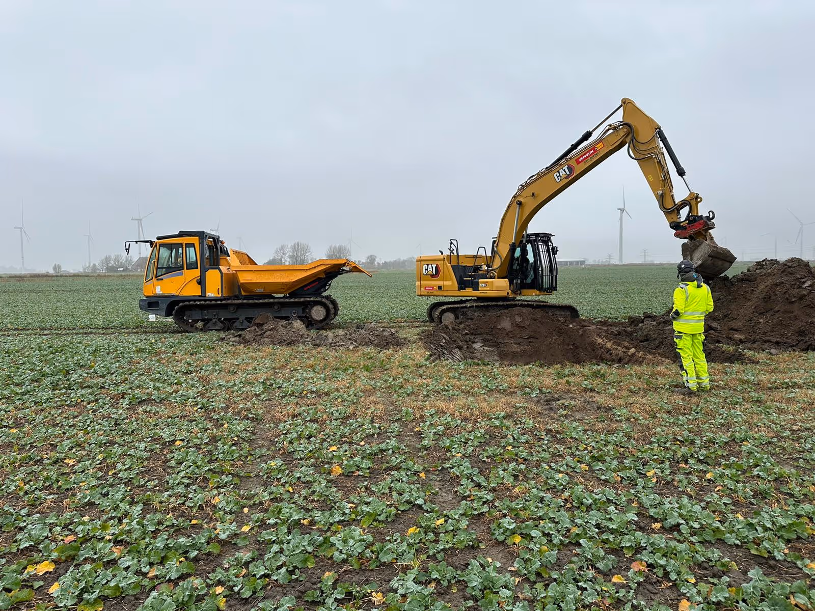 Bauarbeiter in Schutzkleidung steht neben einem Bagger, der Erde in einen Muldenkipper lädt