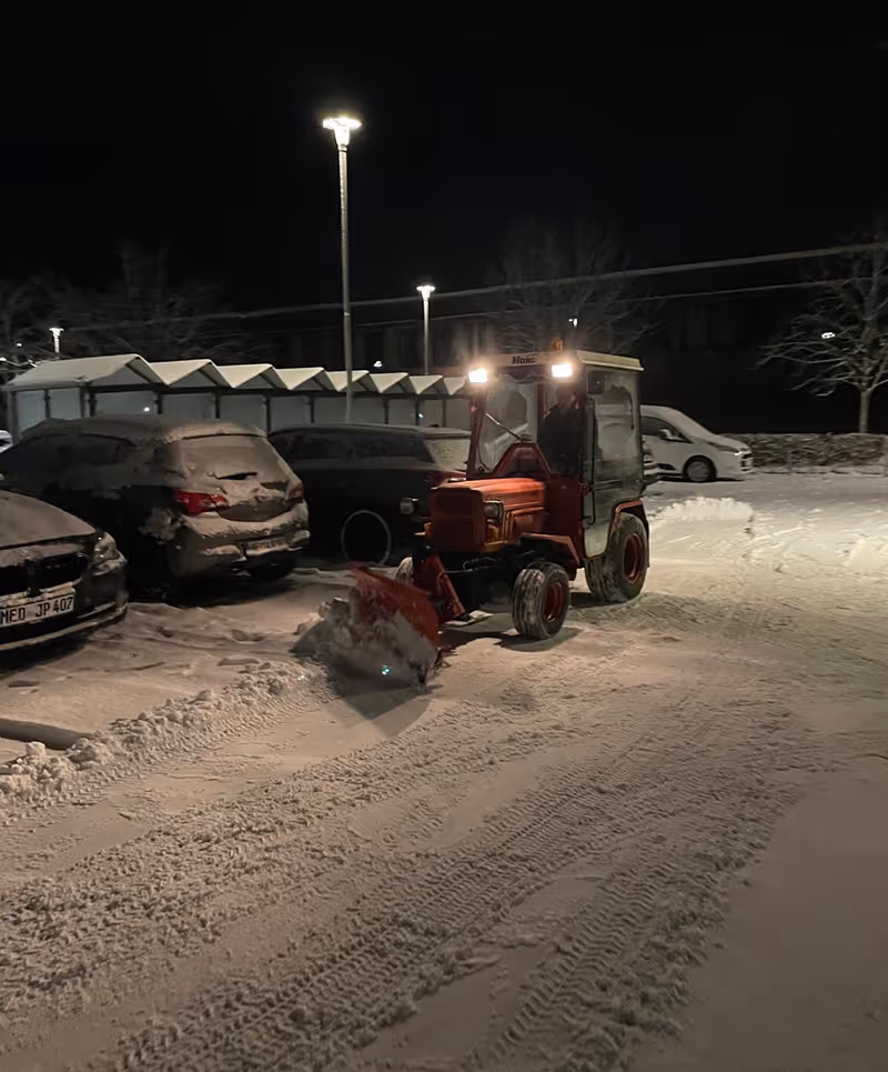 Kleiner roter Traktor mit Schneeschaufel räumt einen schneebedeckten Parkplatz.