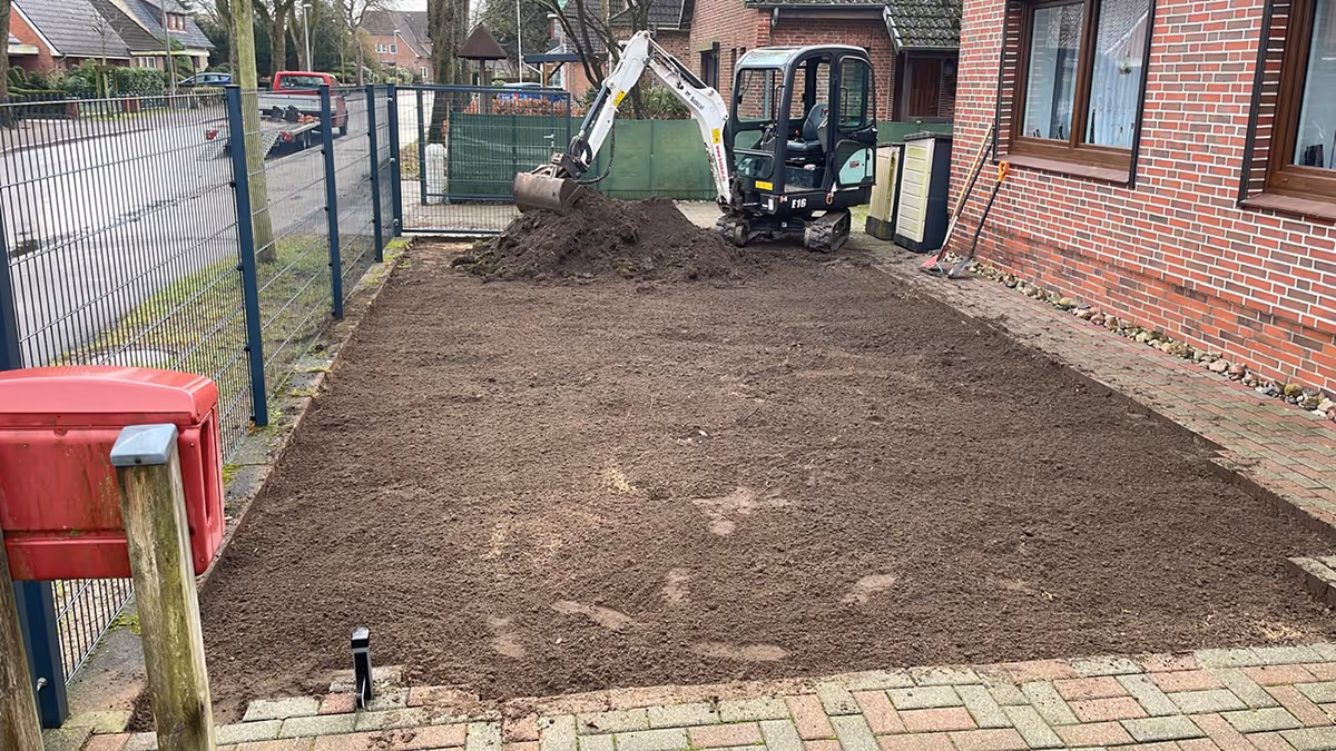 Kleiner Bagger hebt Erde auf einer neu vorbereiteten Fläche neben einem Backsteinhaus.