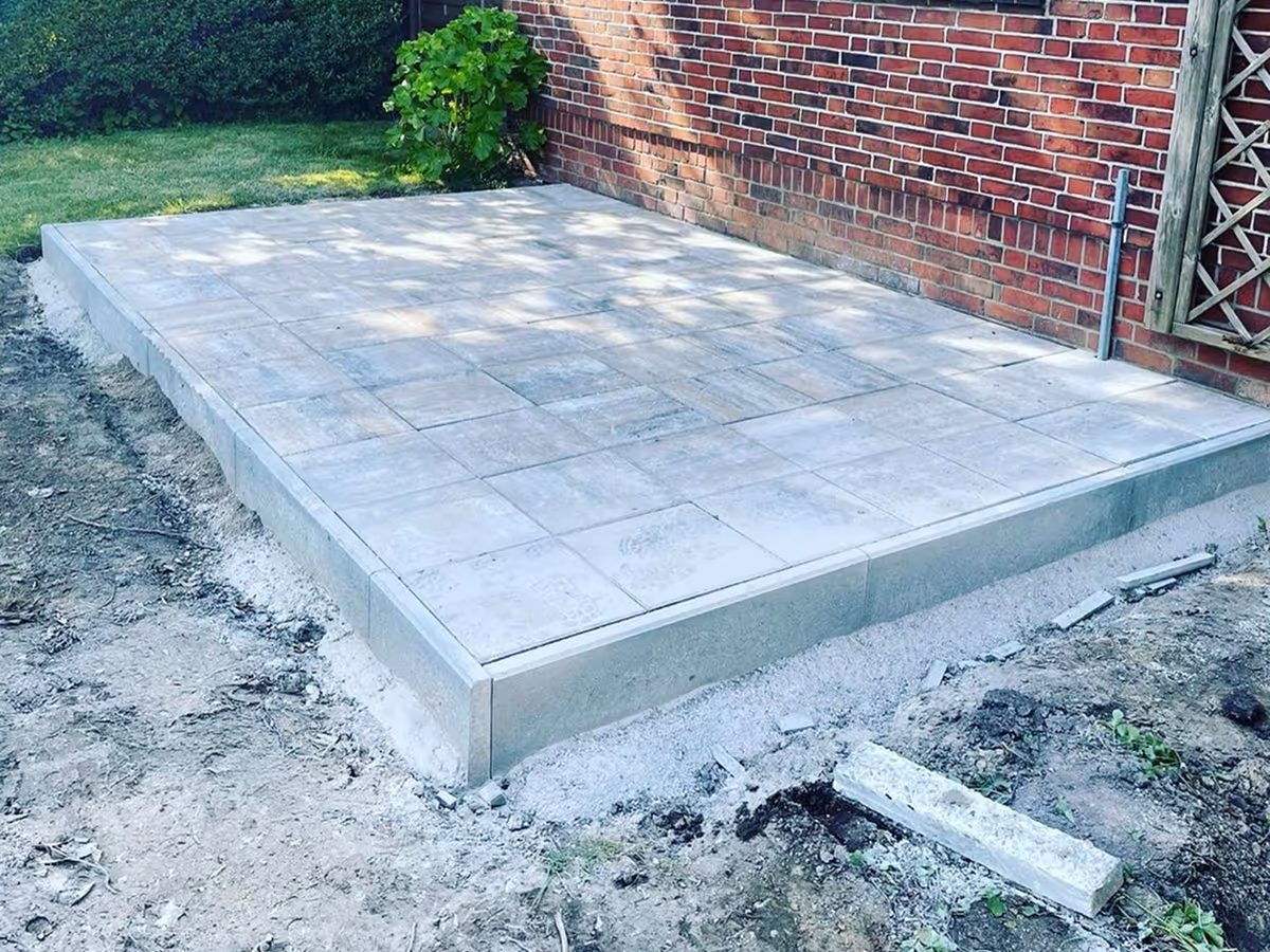 kleine Terrasse mit grauen Betonplatten neben roter Backsteinwand und grünem Garten.