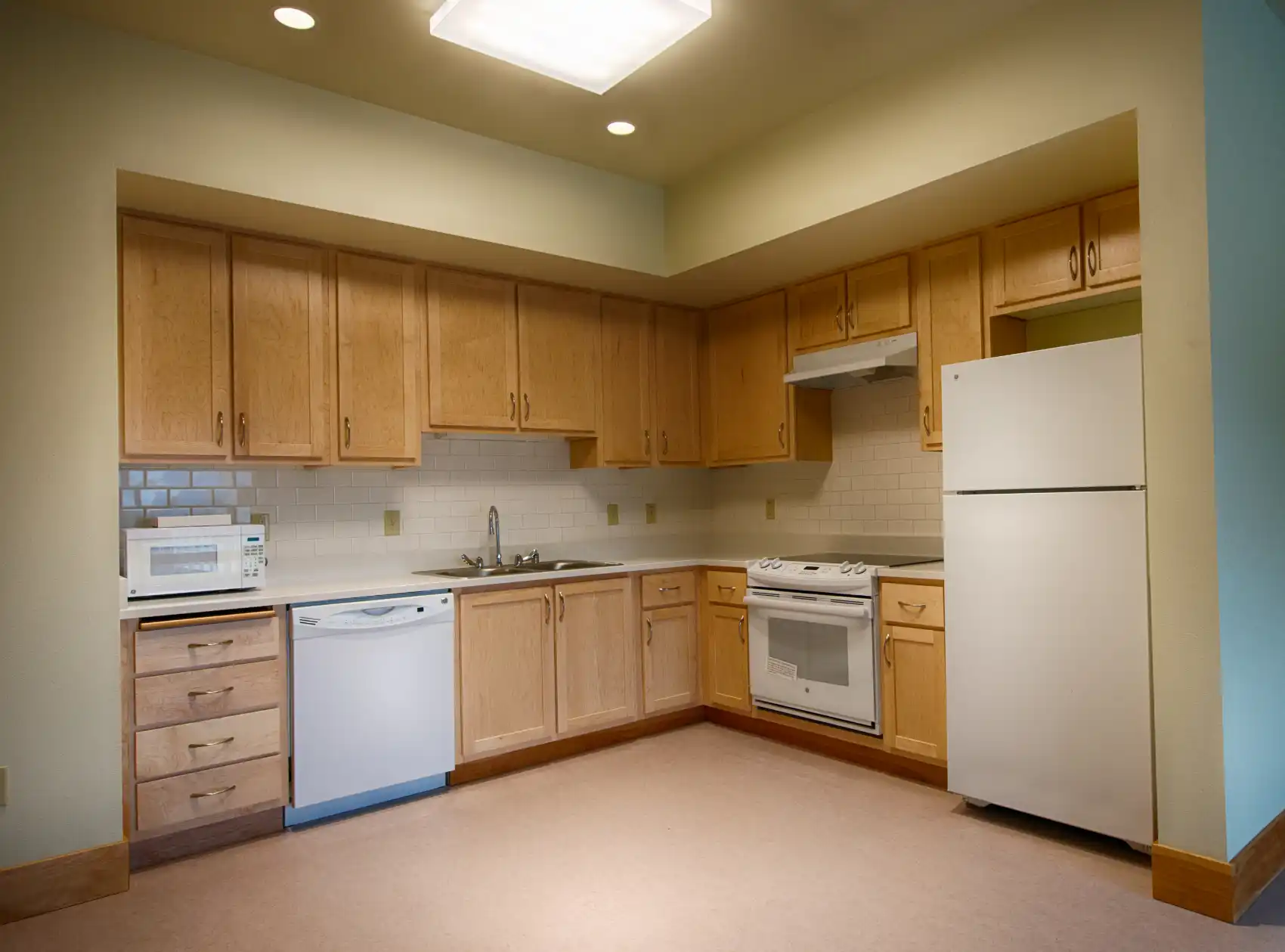 An "L" shaped kitchen with transitional wood cabinets, off-white counter and tile backsplash, and white appliances.