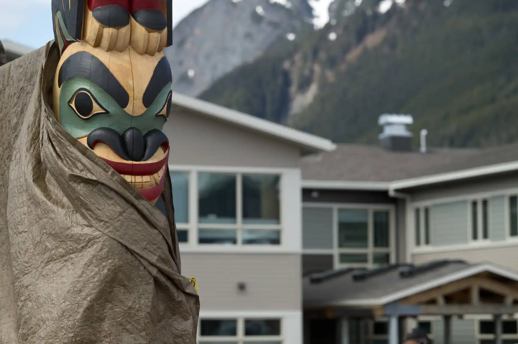 Closeup of a new totem pole, partially wrapped in a protective covering with the new building in the background.