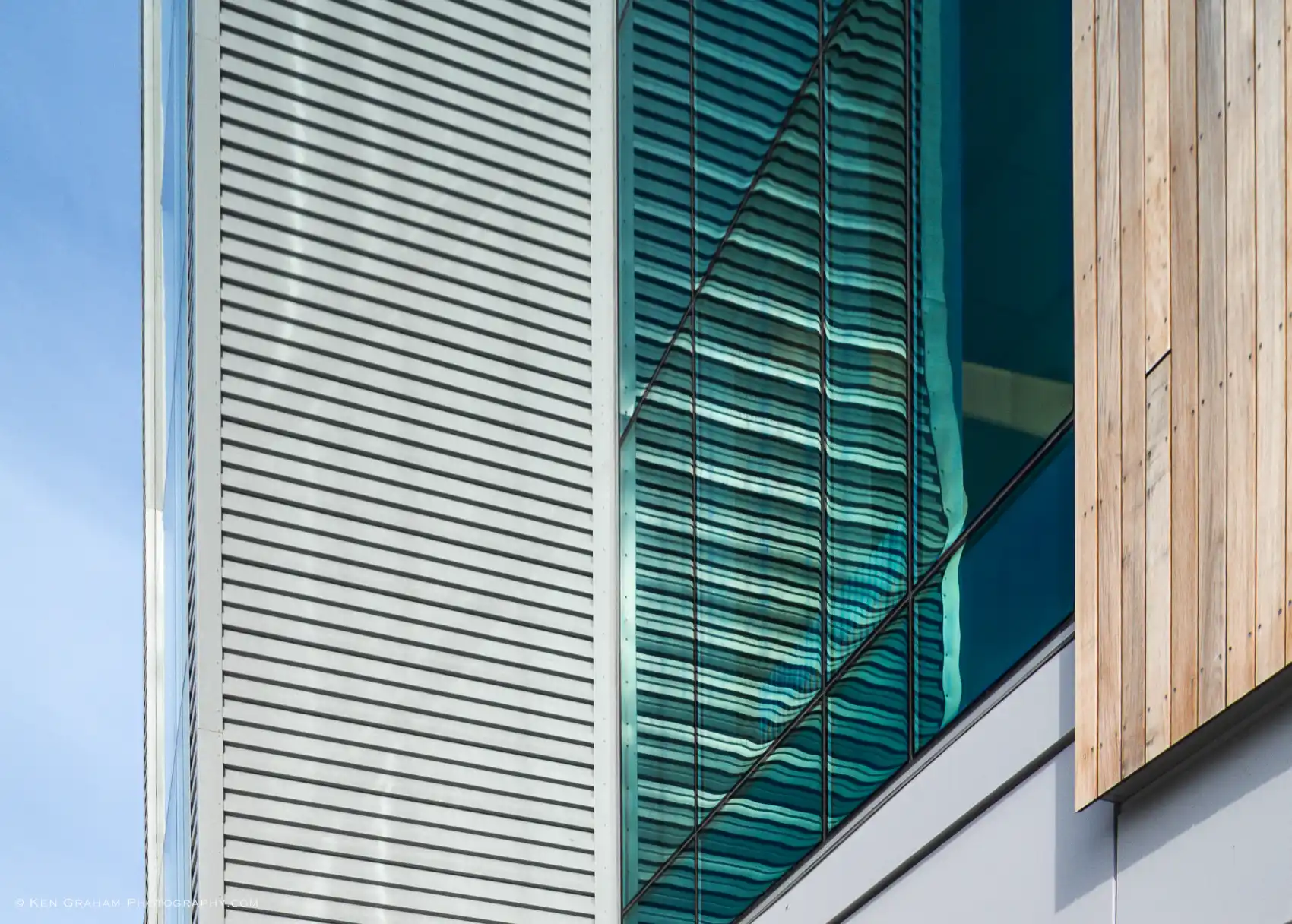 A detail photo of gray corrugated metal siding, blue frameless glass curtain wall, and vertical wood siding.