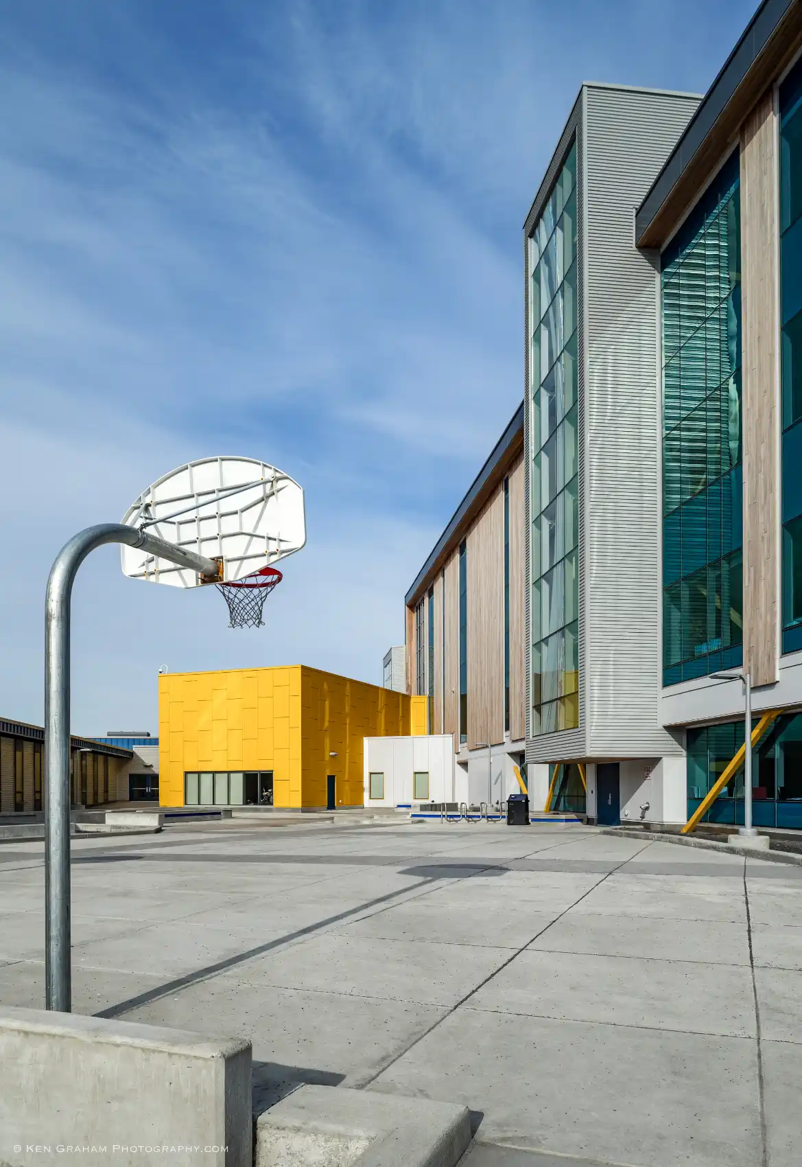 School courtyard with a four-story classroom tower, a smaller yellow volume as the band rooms, and concrete paving, seating, and basketball hoop.