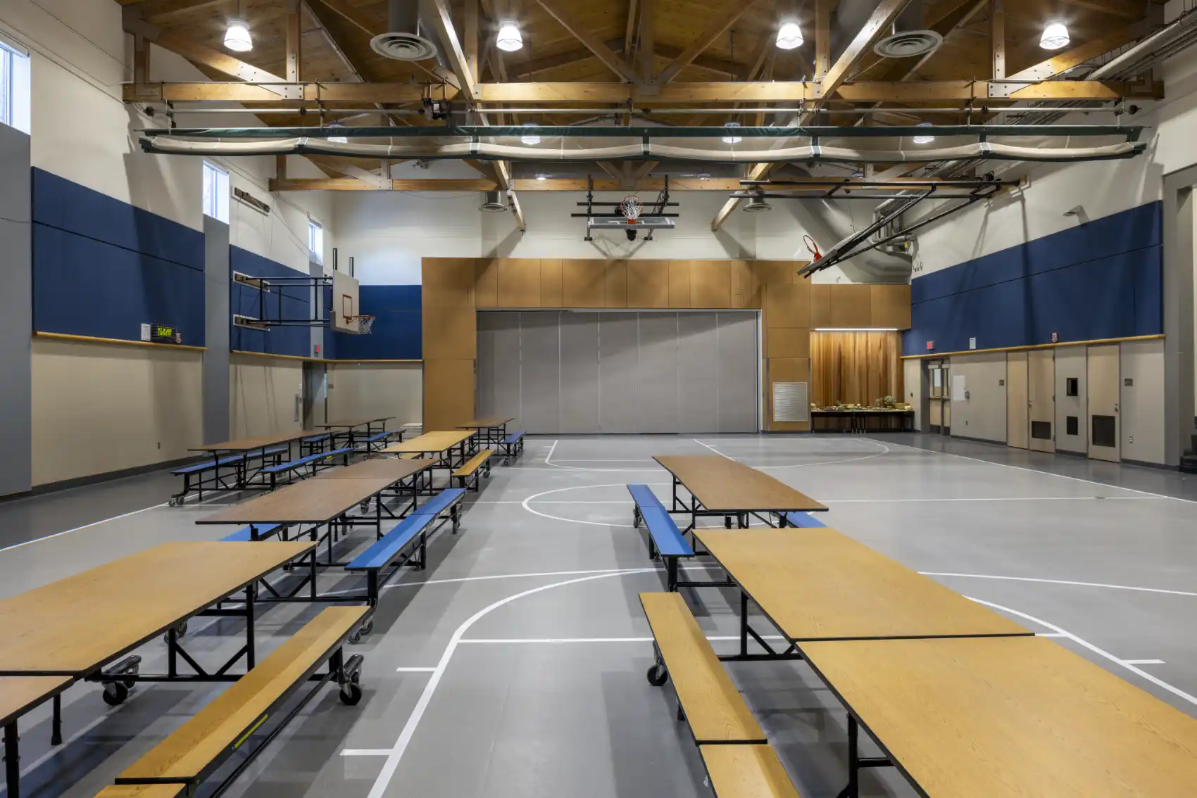 Multi-purpose room with a gray court floor, painted and wood paneling, blue acoustical panels, and wood ceiling and trusses.