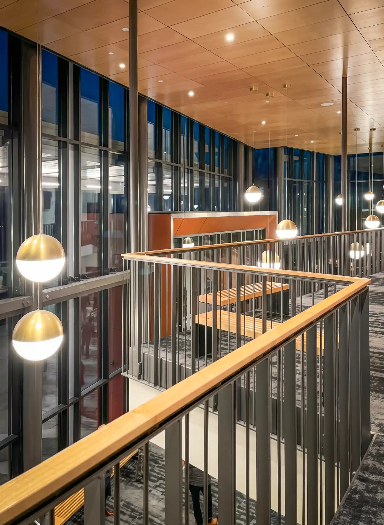 View of the lobby from the balcony with dark metal railing with a wood handrail, wood ceiling, and brass globe hanging lights.