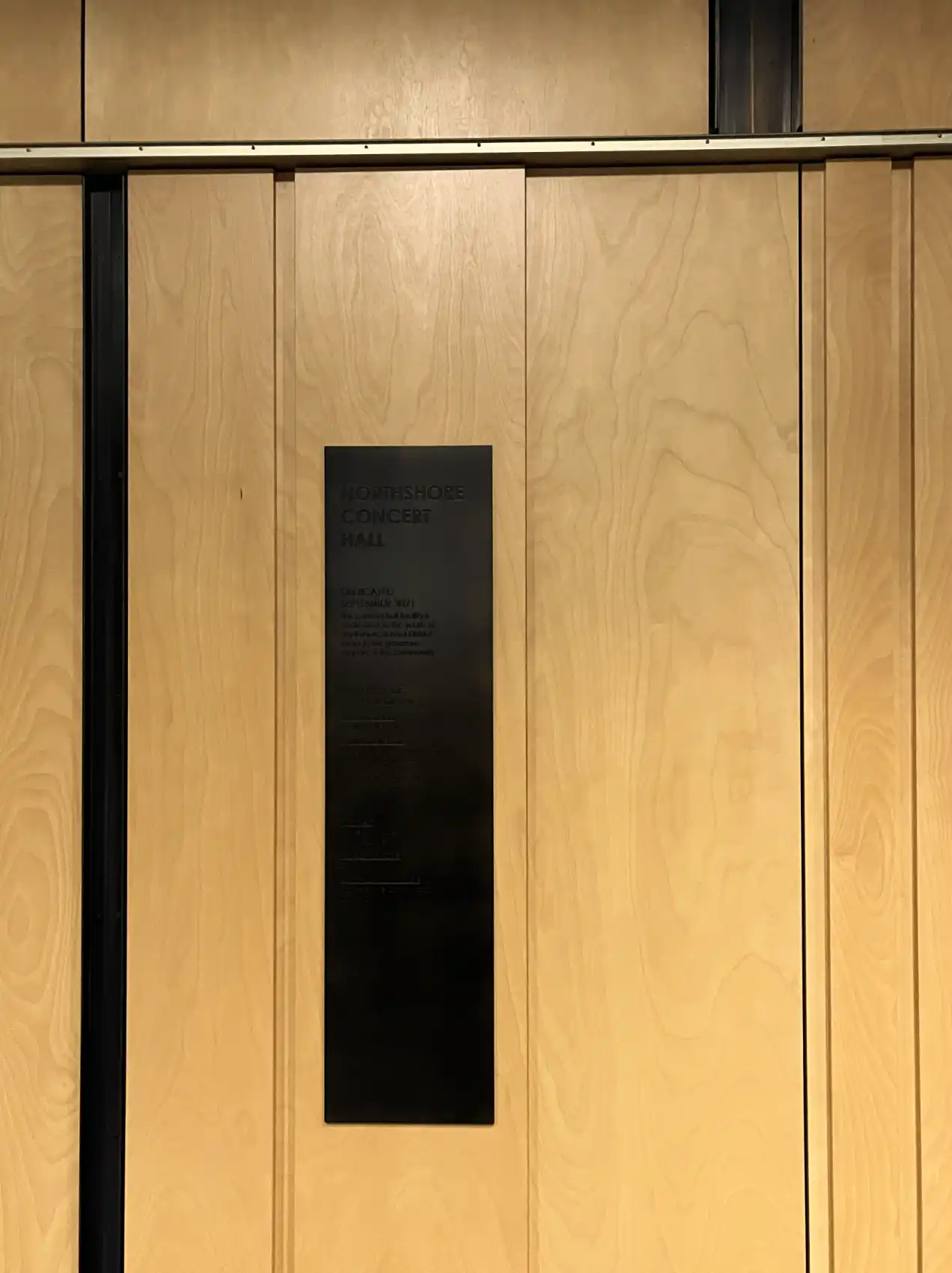 Light wood paneling with brass and dark bronze metal accent strips; dark bronze dedication plaque.