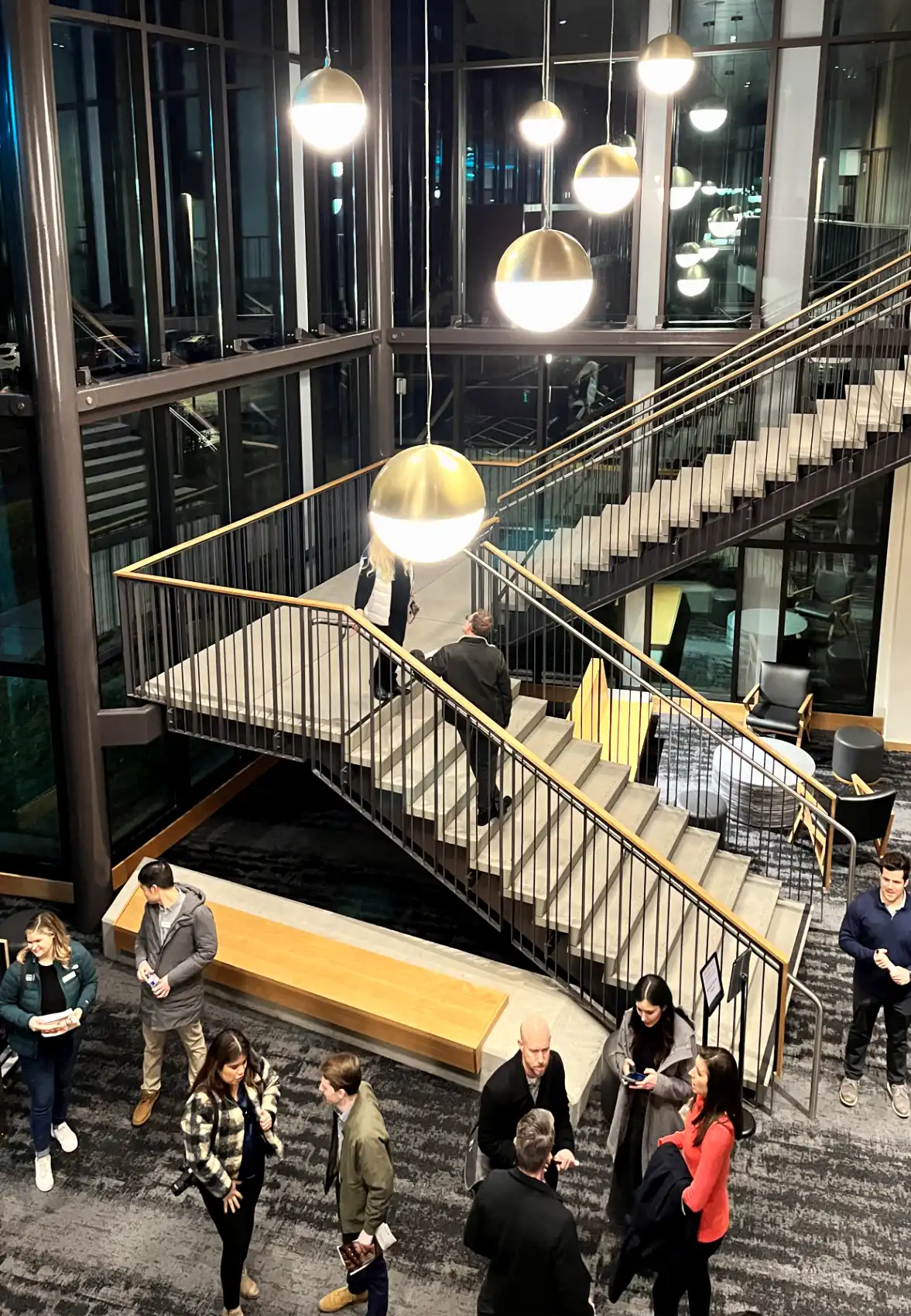 Modern floating concrete stairs with metal and wood railing with concrete and wood benches at the bottom, surrounded by people.