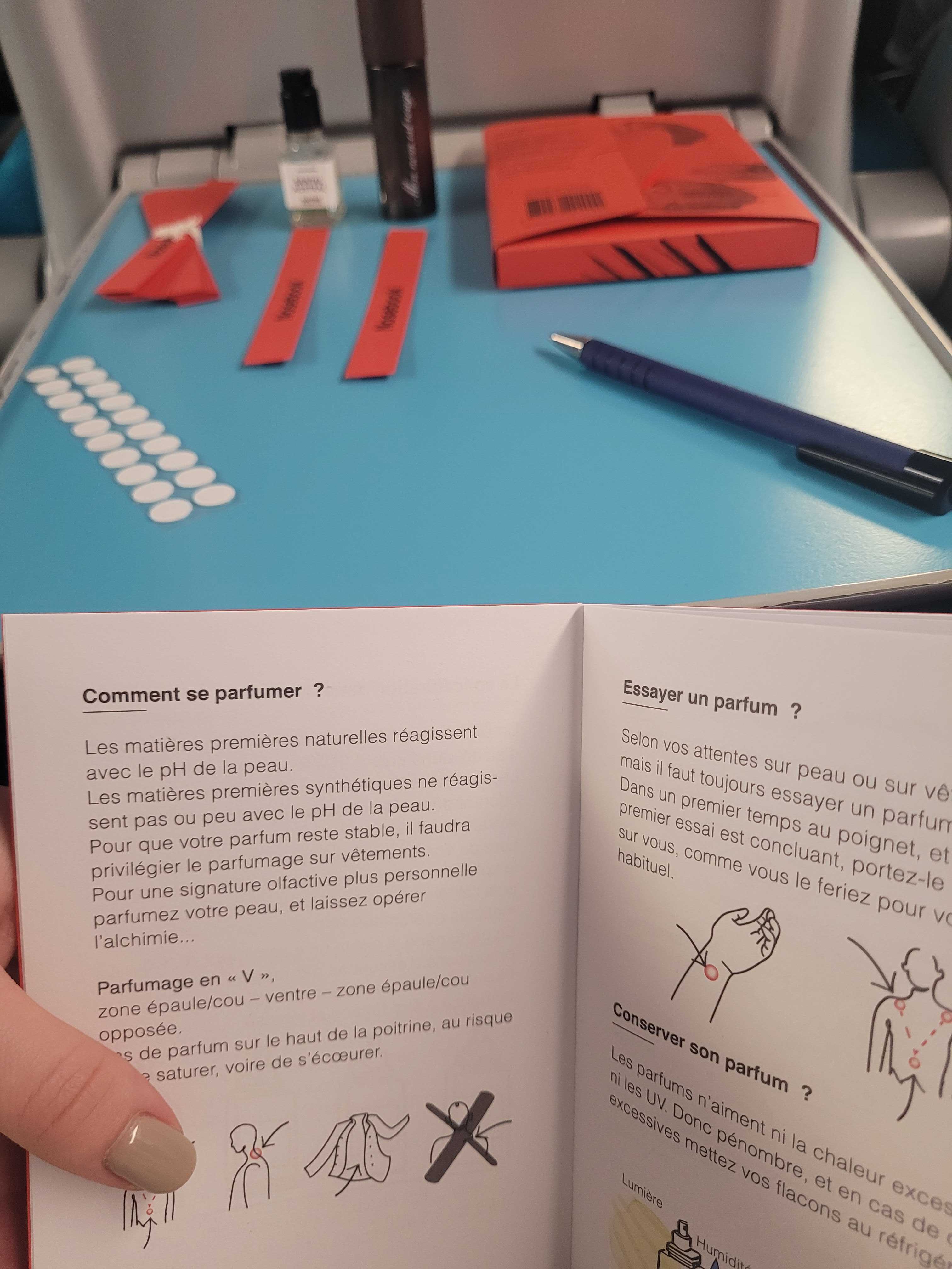 Set de parfumerie avec 10 petits échantillons, un coffret rouge "Nosebook" ouvert avec un stylo, une touche rouge et une tasse blanche sur une table noire.