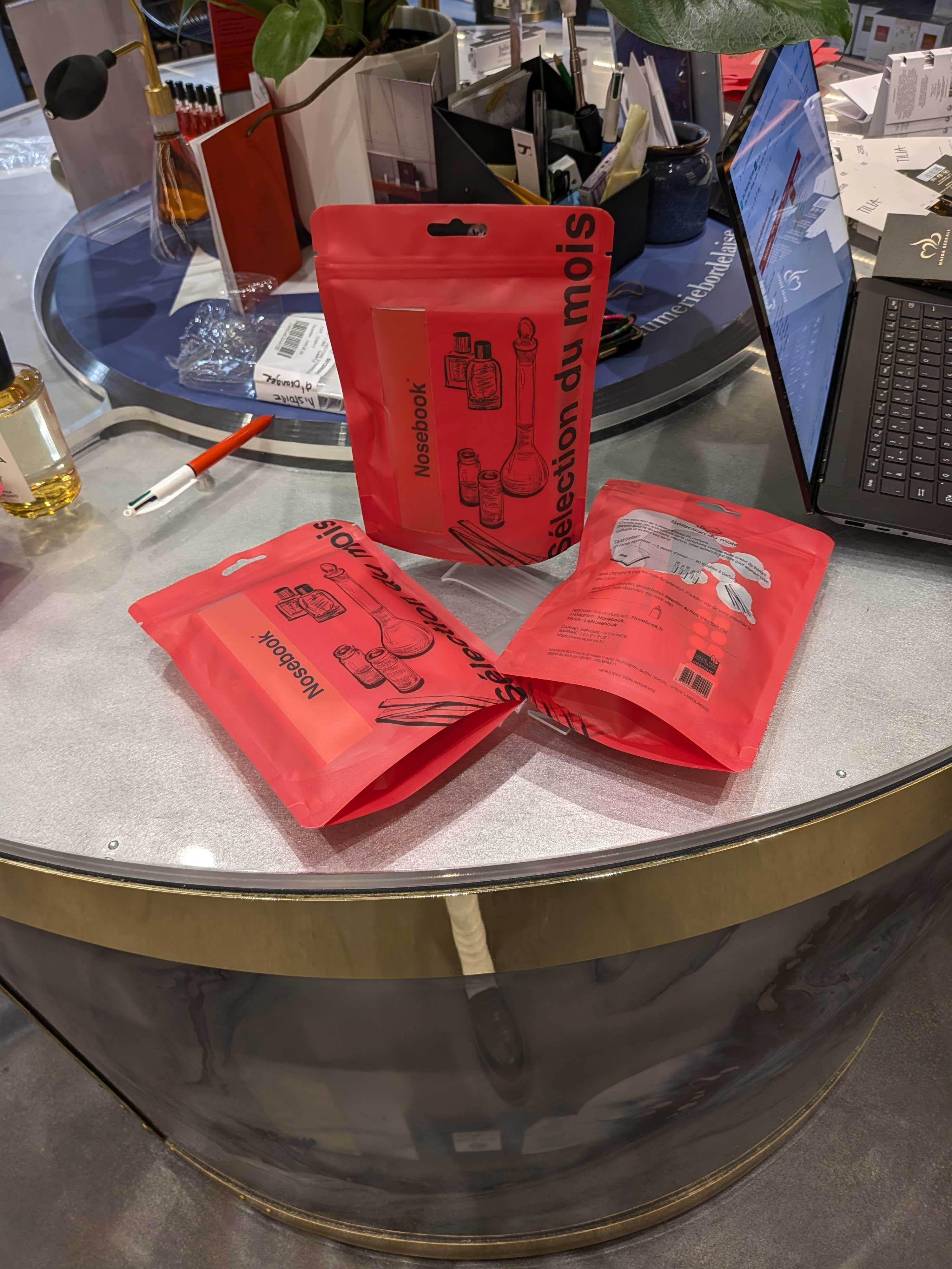 Main droite tenant un carnet rouge avec l'inscription noire "Nosebook" et un sceau embossé, made in France au tour et merci au centre.