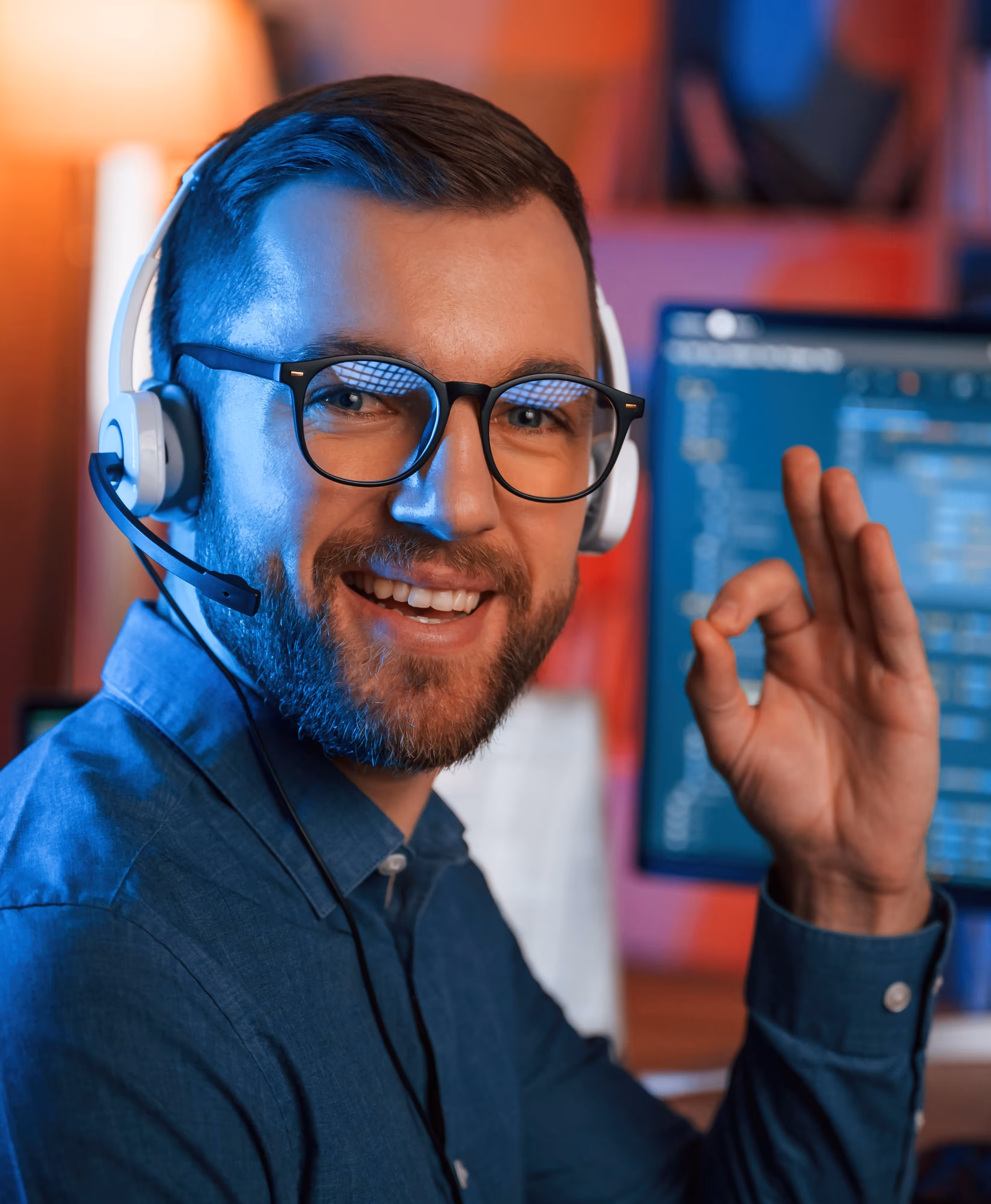 Smiling man wearing headset and glasses showing OK hand sign while sitting in front of a computer with code on the screen.
