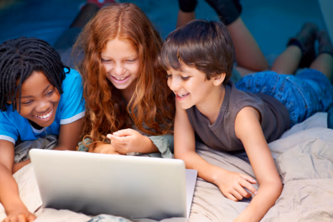 Three children lying on a bed, smiling and looking at a laptop screen together.