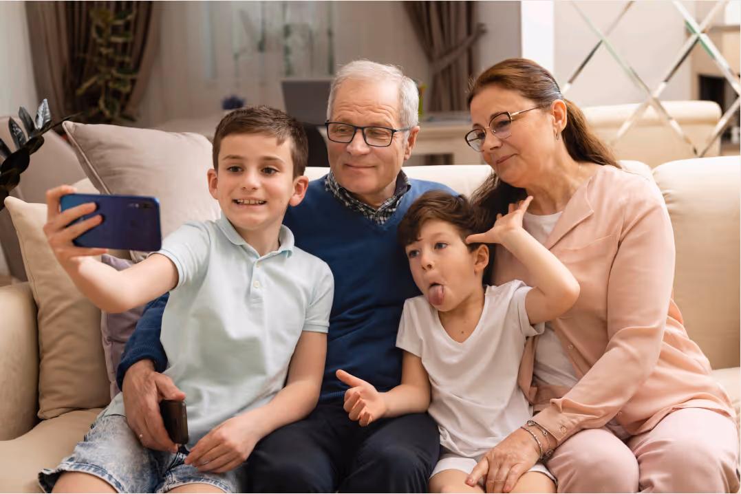 Two boys and an elderly couple sitting on a couch, one boy taking a selfie while the other makes a funny face.