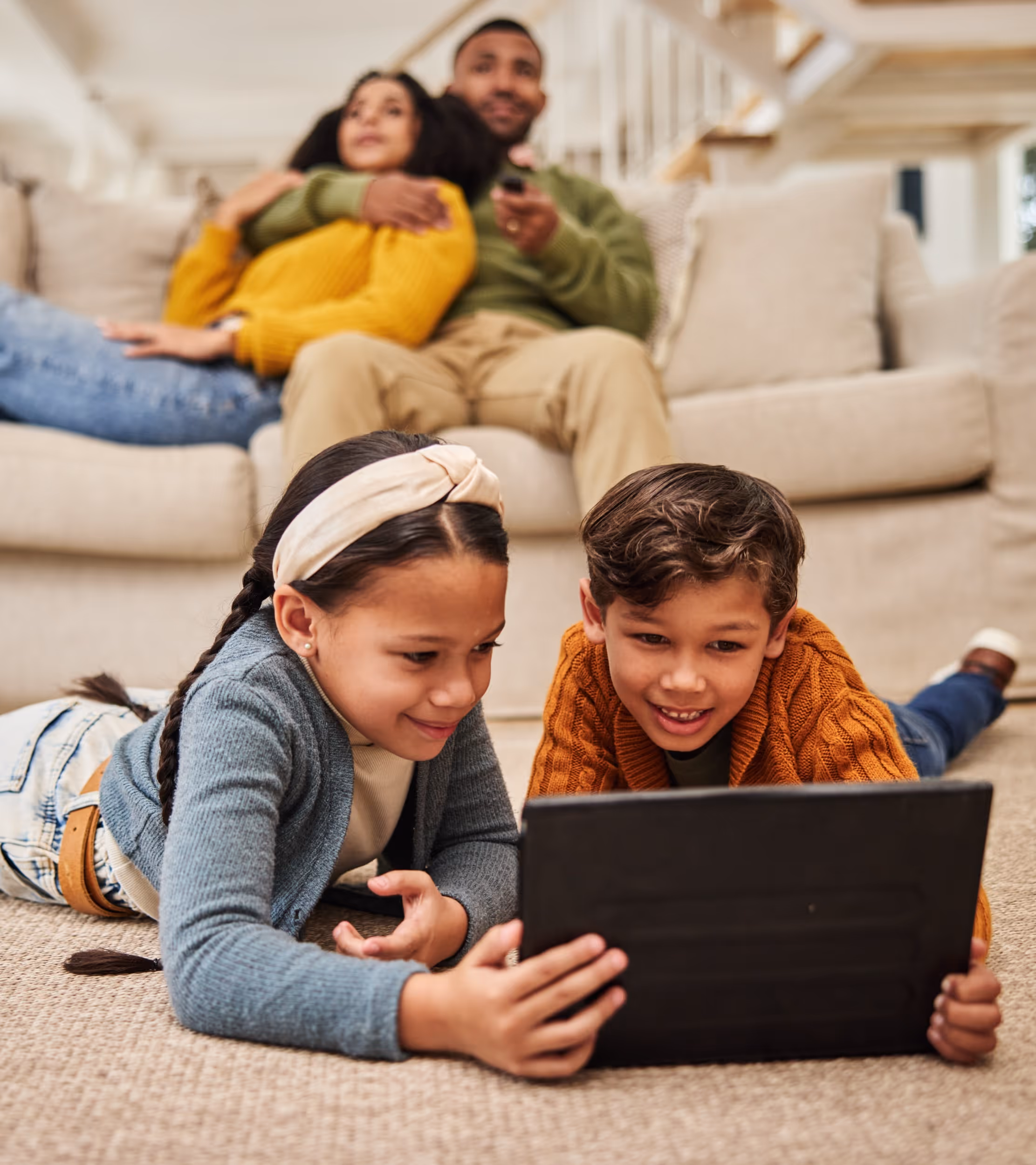 Two children lying on a carpeted floor watching a tablet, with a couple sitting on a couch in the background.