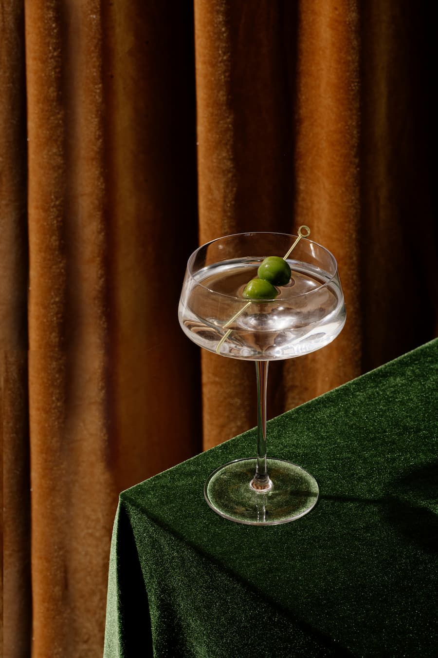 A martini glass with two green olives on a skewer, placed on a green velvet table with brown drapes behind.