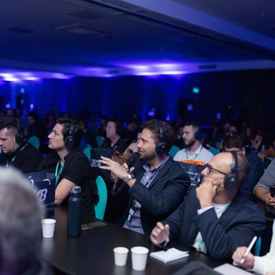 Participantes da plateia usando fones de ouvido, atentos a uma palestra em um evento, com iluminação ambiente azul.
