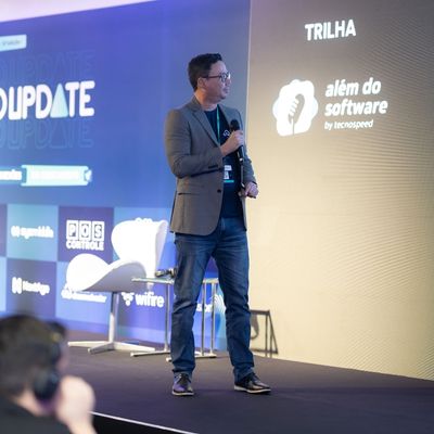 Homem usando blazer e jeans, segurando um microfone e falando no palco, em frente a uma tela com o logo “Trilha Além do Software by TecnoSpeed”.