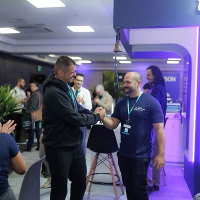 Dois homens apertando as mãos e sorrindo em um evento interno movimentado, com outras pessoas ao fundo.