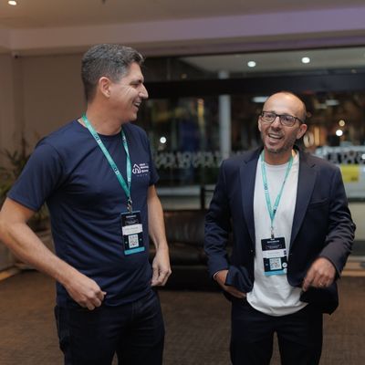 Dois homens usando crachás de evento, sorrindo e conversando em um ambiente interno.