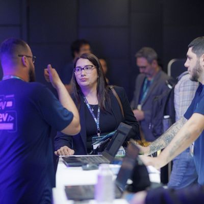 Mulher usando óculos e cordão, conversando com dois homens em uma mesa de registro ou informações em um ambiente de conferência.