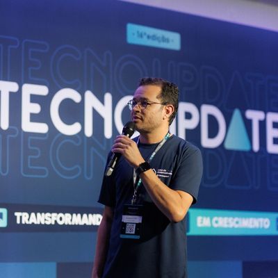 Homem usando óculos, falando ao microfone no palco, em frente a um banner com o nome TECNOUPDATE.